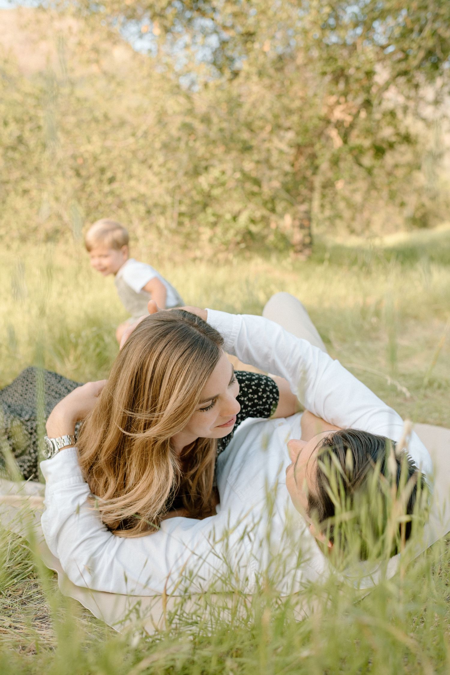 Couple lying on a blanket in a grassy field sharing a tender moment while child plays behind them.