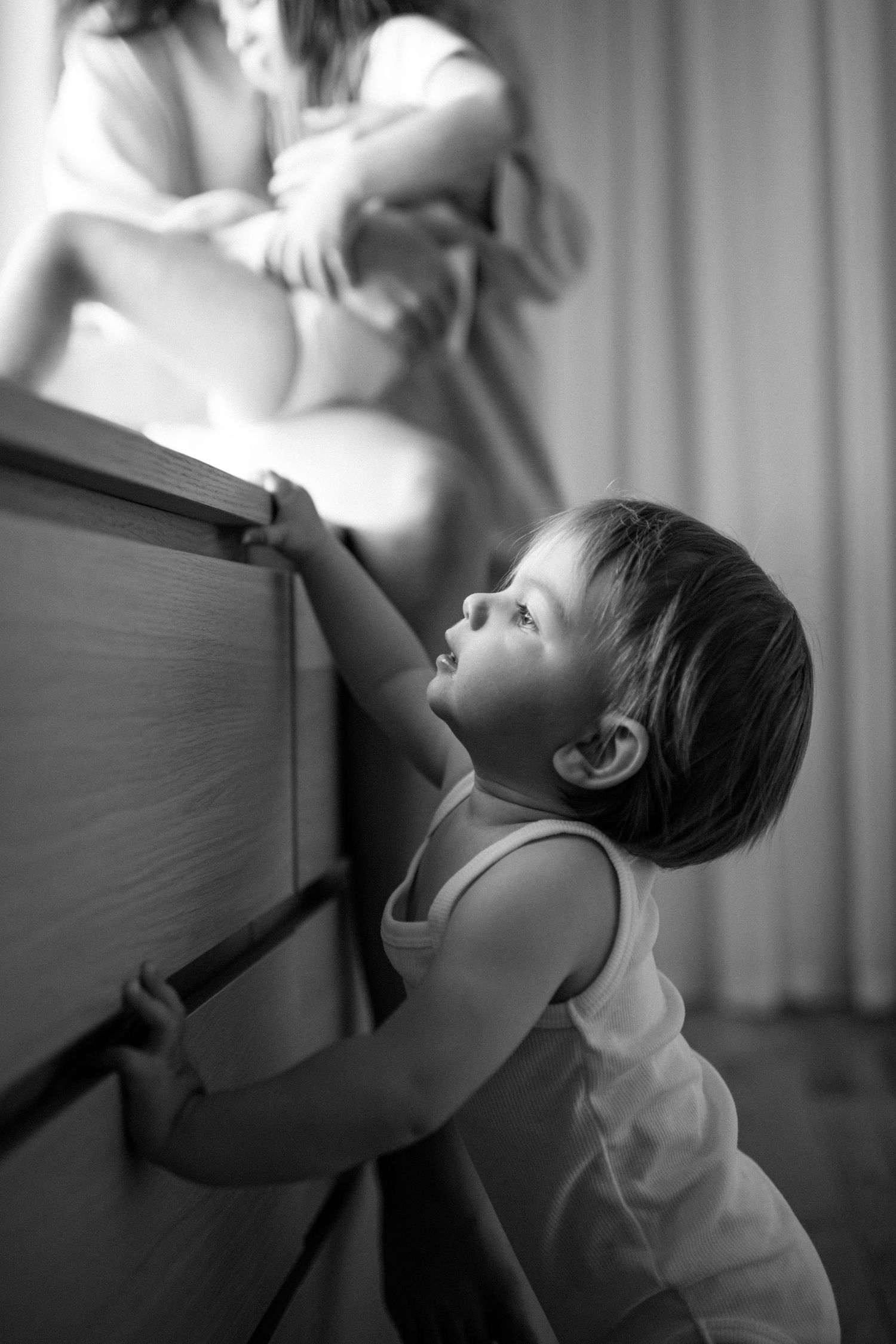 Black and white photo of a toddler in a tank top reaching up to grab the edge of a wooden dresser.