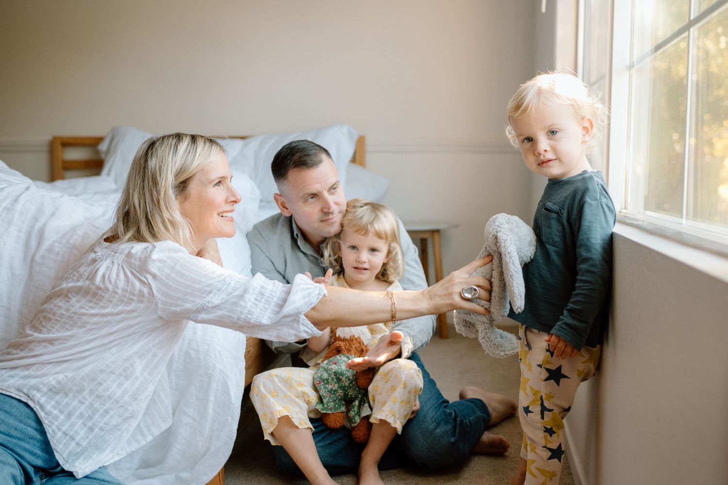 Happy family of four sitting together near a window as mother reaches toward standing toddler.