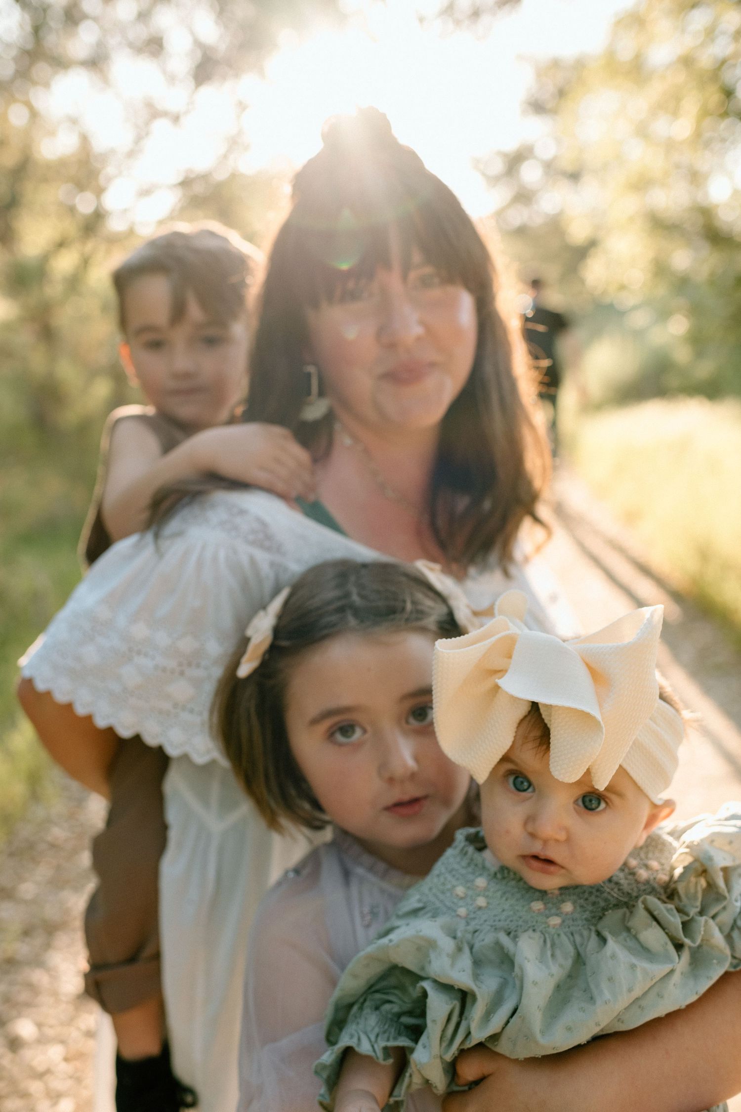 Mother with dark bangs holding three young children outdoors in golden sunlight through trees.