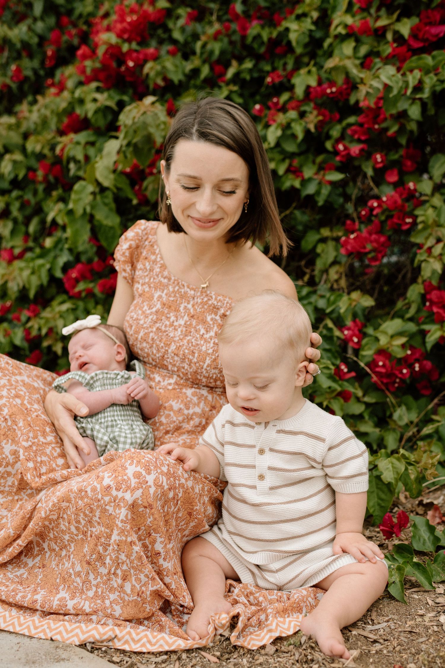 Mother in floral dress sitting outdoors with newborn and toddler in front of red flowering bushes.