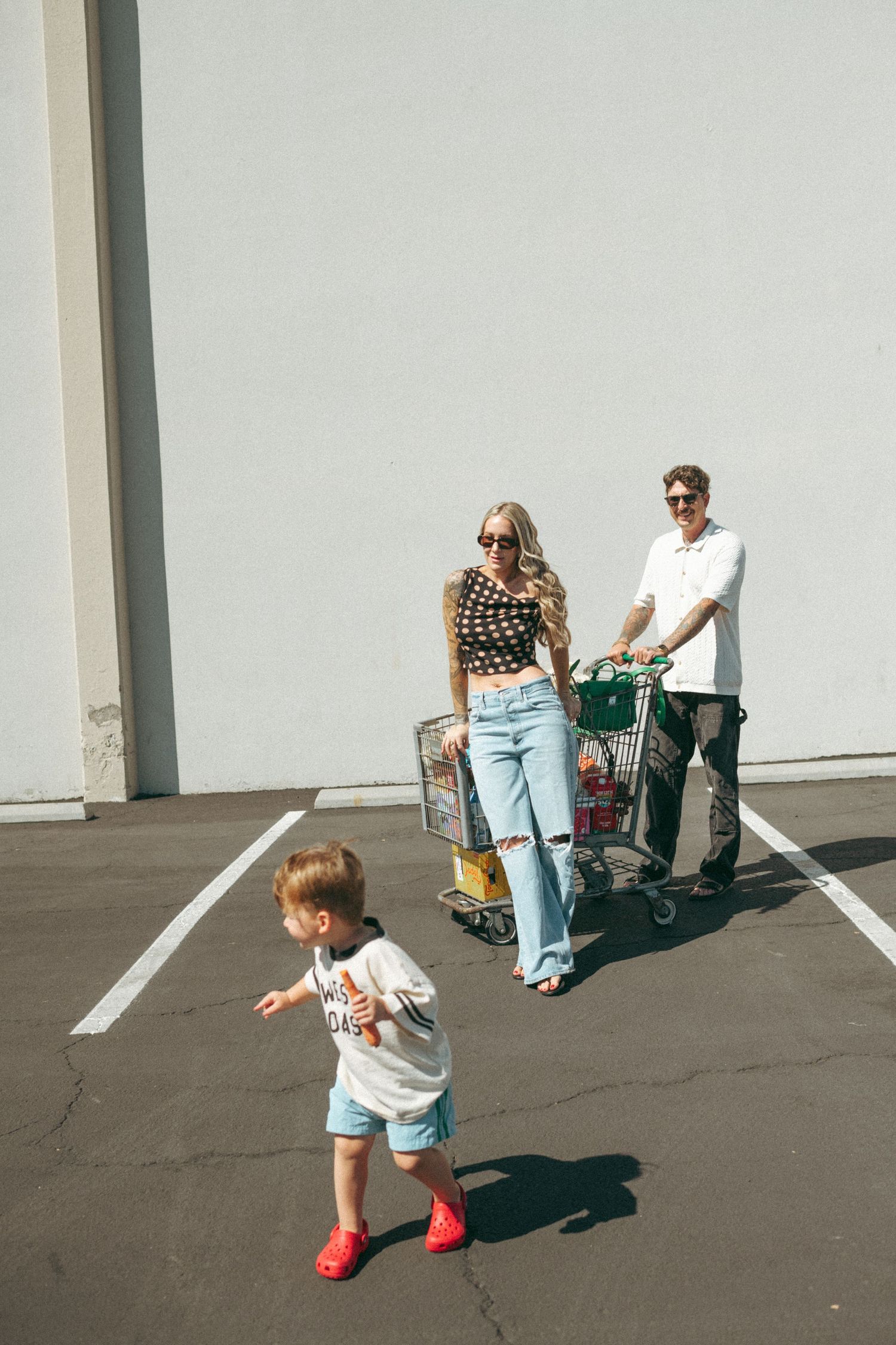 Family with shopping cart in sunny parking lot, mom in polka dot top and jeans, dad in white shirt, toddler in red shoes.