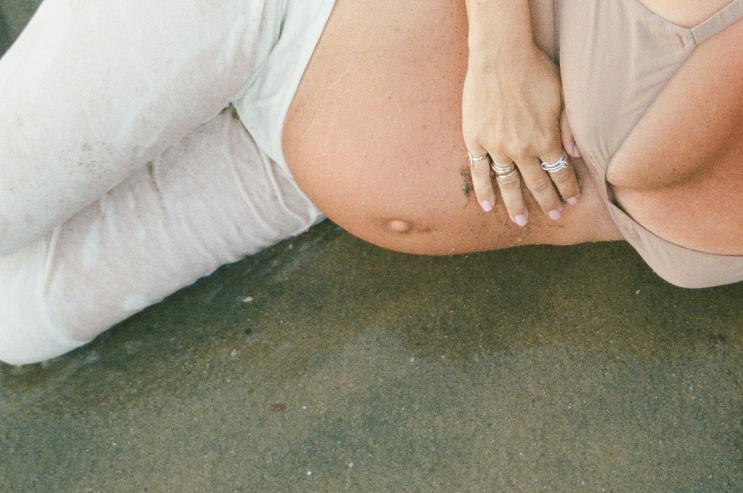 Close-up of a pregnant woman's bare belly at the beach, wearing rings and light-colored clothing.