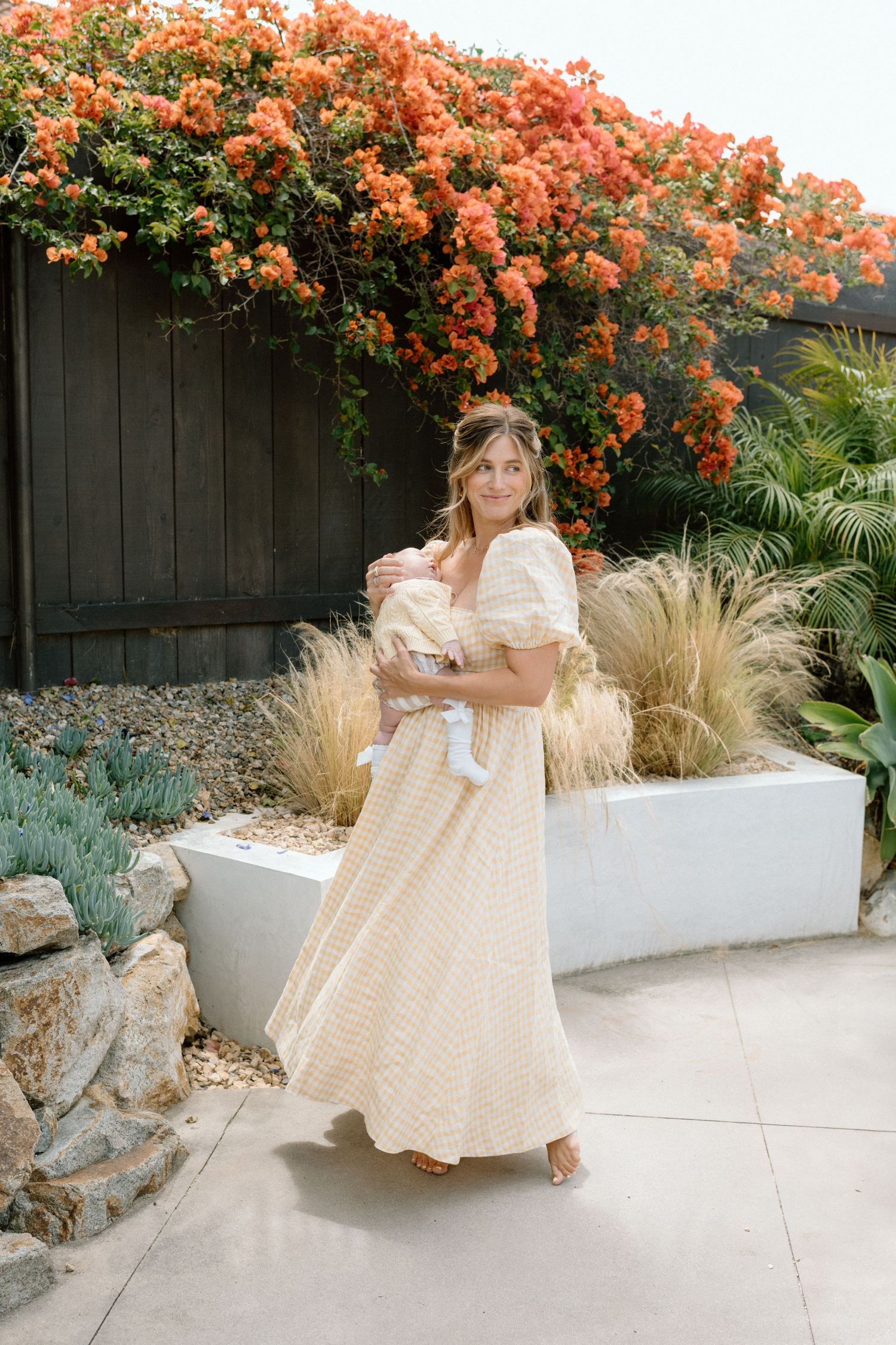 Mother in yellow gingham maxi dress holding newborn baby outdoors with bougainvillea.