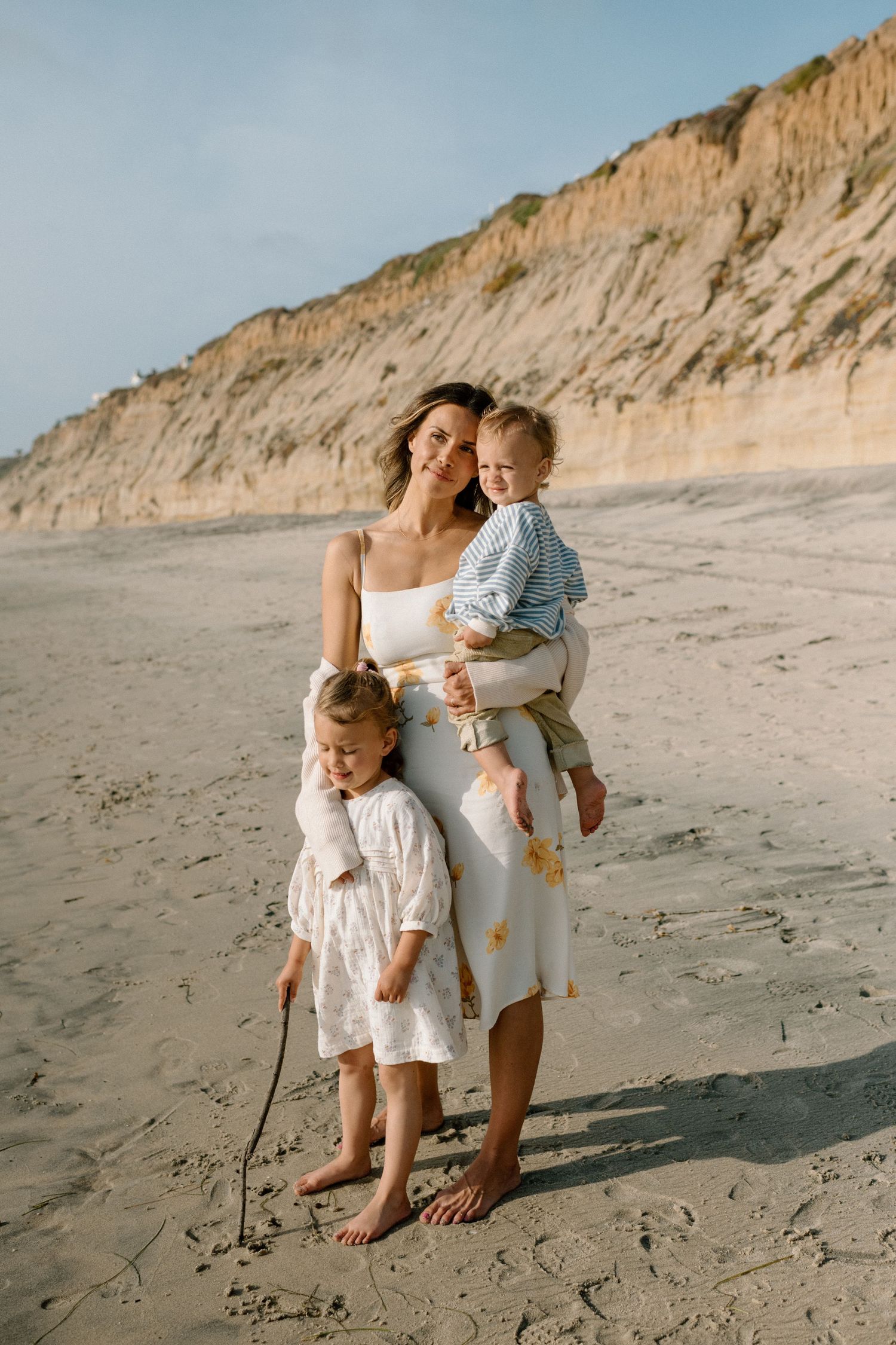 A mother holds her baby while her toddler stands beside her on a sandy beach with cliffs in the background.