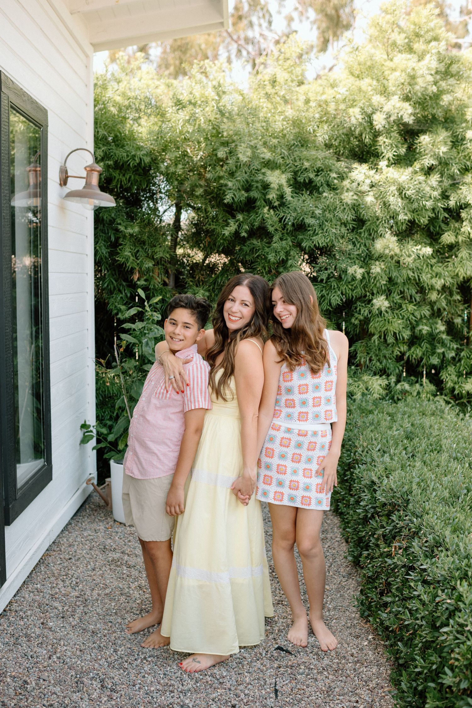 Three people in summer outfits stand together on a pebbled patio against lush green foliage and white house exterior.