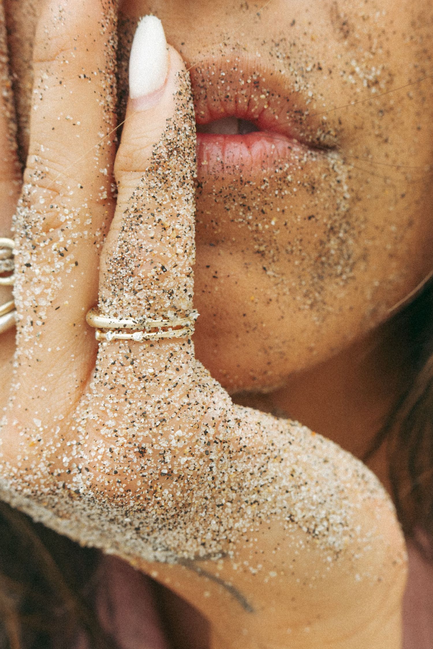 Close-up of woman's face and hand covered in beach sand wearing gold stacked rings.