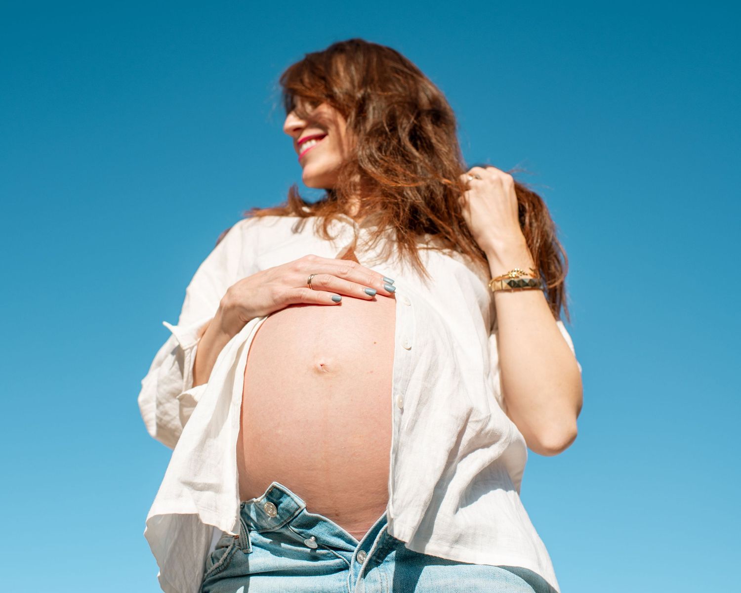 Smiling pregnant woman in open white shirt and jeans showing baby bump against blue sky.