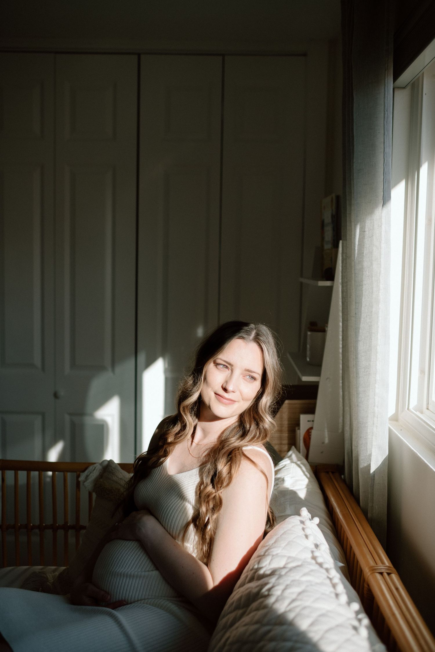 Pregnant woman with long wavy hair sitting peacefully by a sunlit bedroom window.
