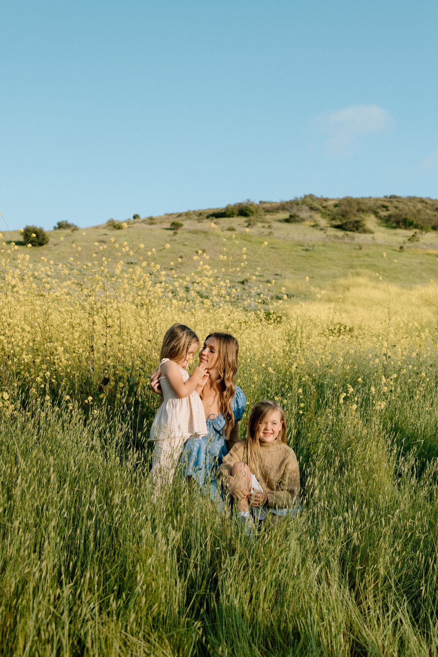 A mother sits in a wildflower field with her two young daughters on a sunny day.