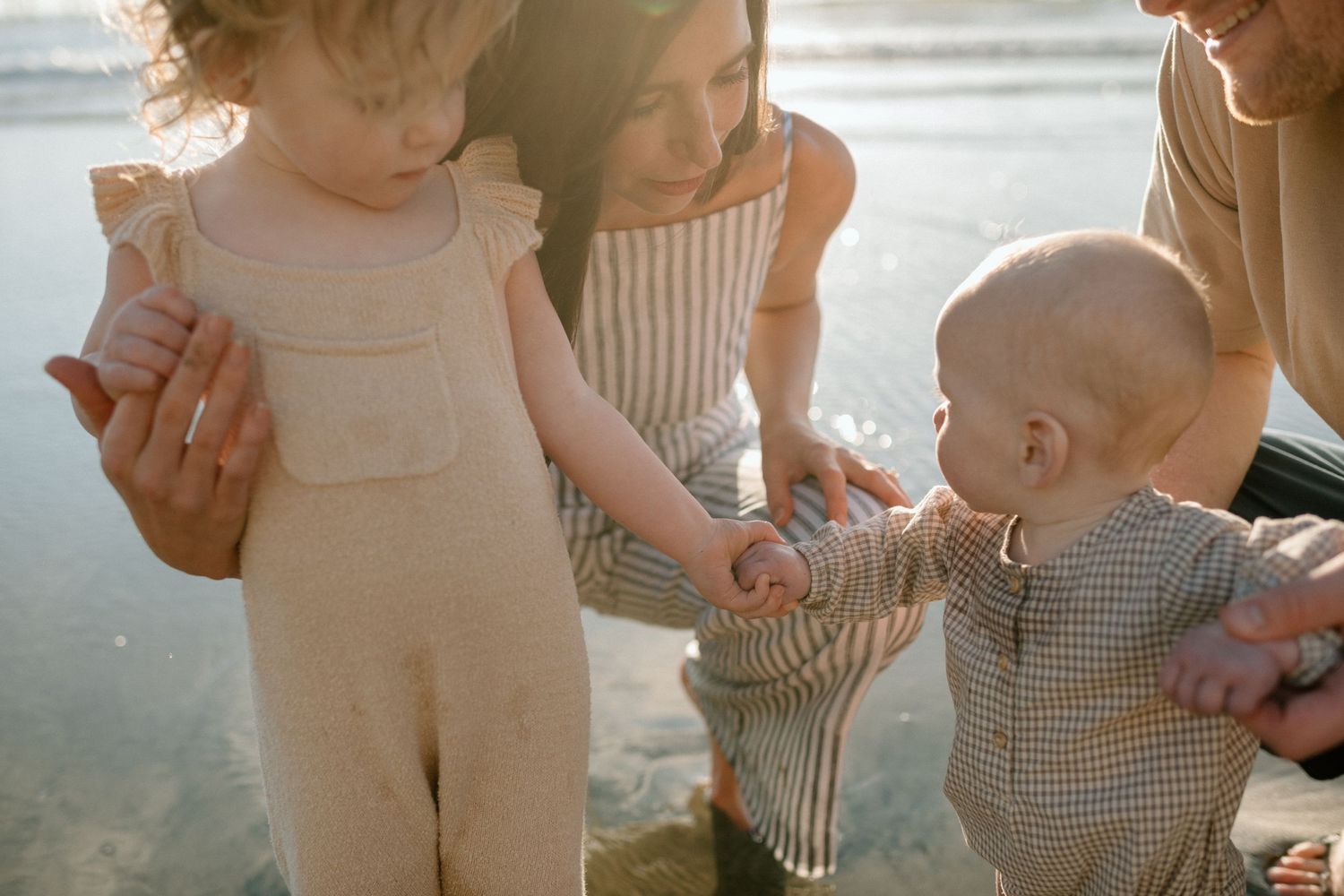 Young toddler in linen romper touching baby sibling's hands while mother watches at beach.