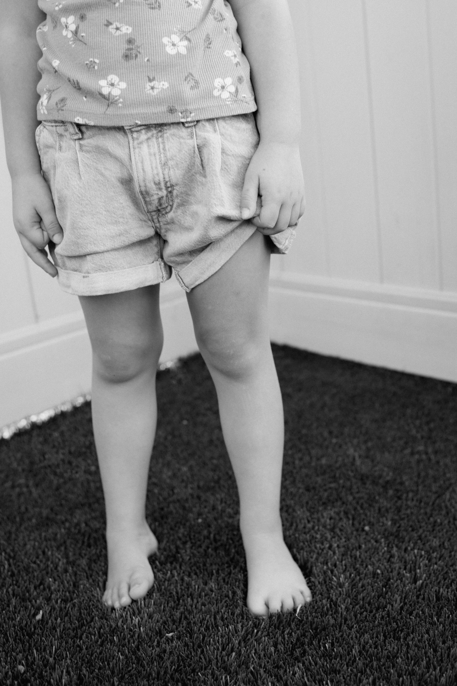 Black and white close-up of young barefoot girl in denim shorts and floral top standing on grass.