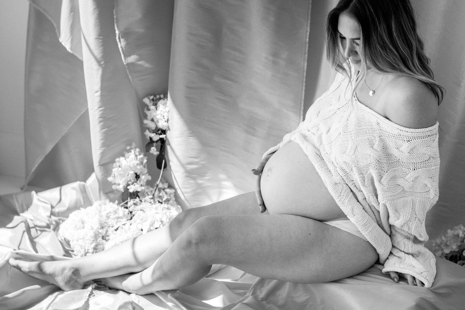Black and white maternity photo of pregnant woman in white lace robe sitting gracefully with flowers.