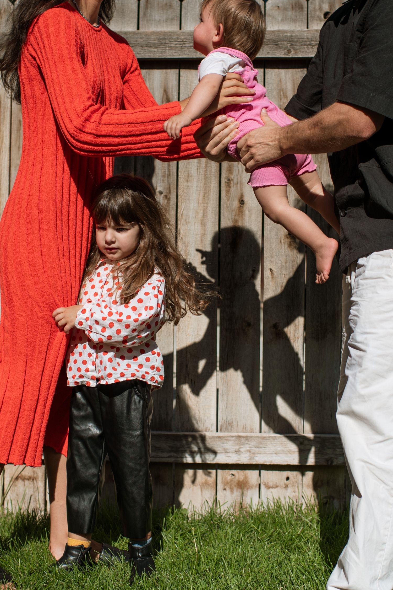 Family outdoors by wooden fence, mother in red dress and father holding baby in pink, young girl standing nearby.