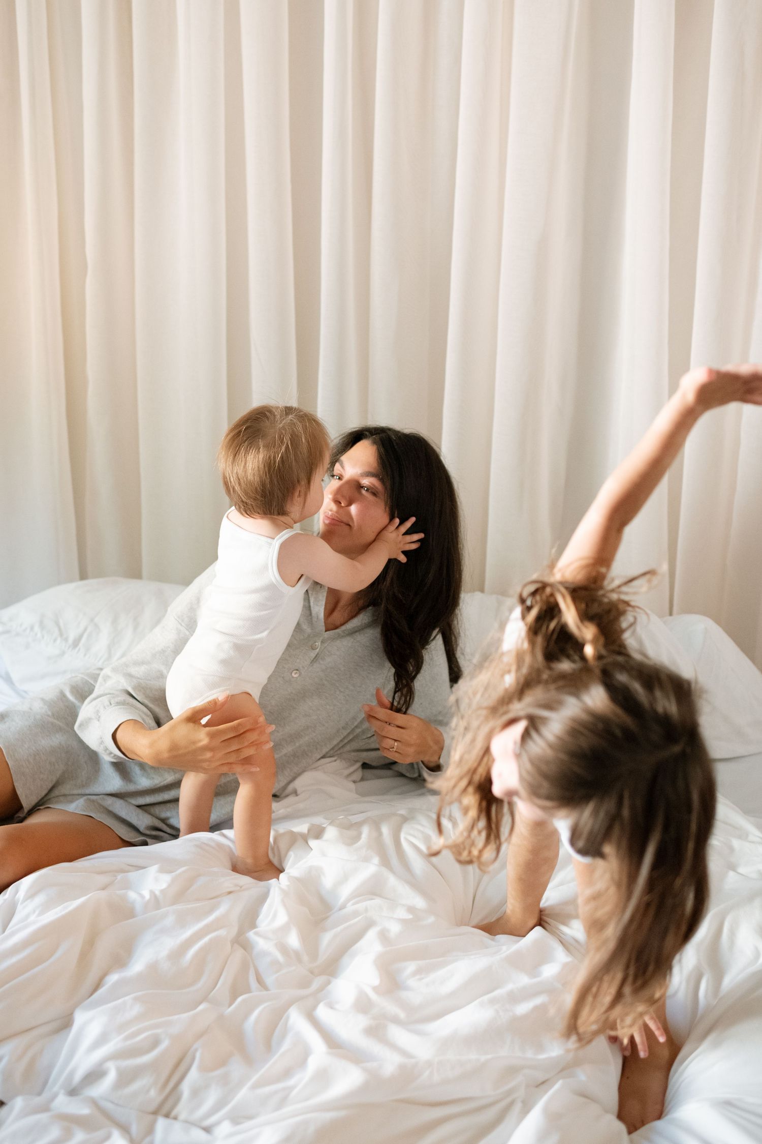 A mother sits on a white bed, holding her baby while an older child plays joyfully beside them in a bright room.
