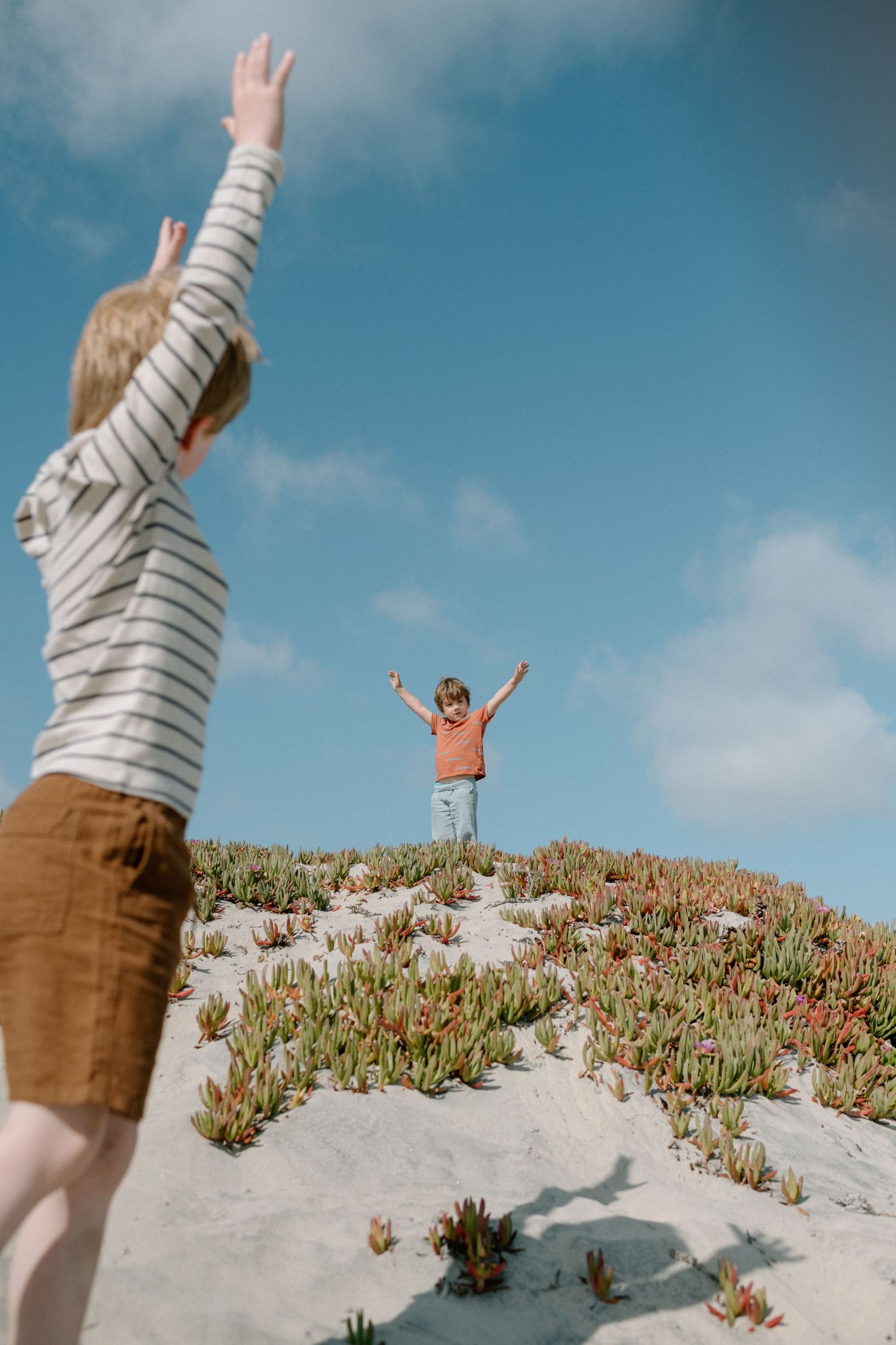 Two young children raise their arms joyfully on a sandy dune covered with coastal plants under a blue sky.