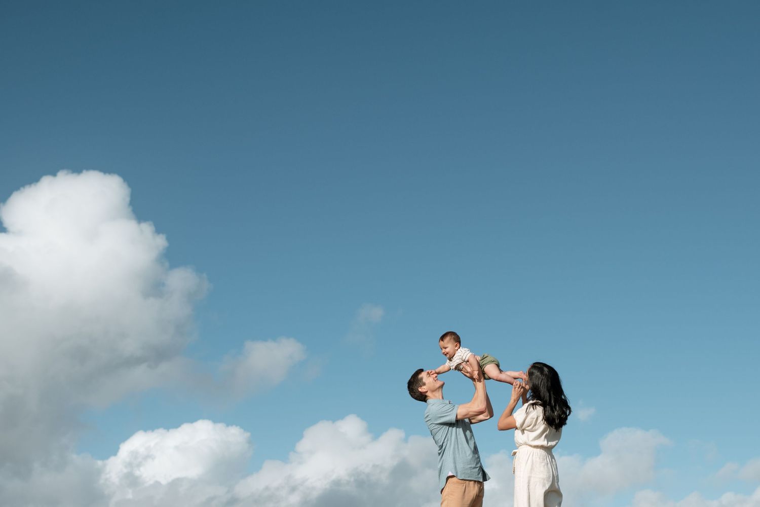 Parents lifting smiling baby into a blue sky with white clouds on a bright sunny day.