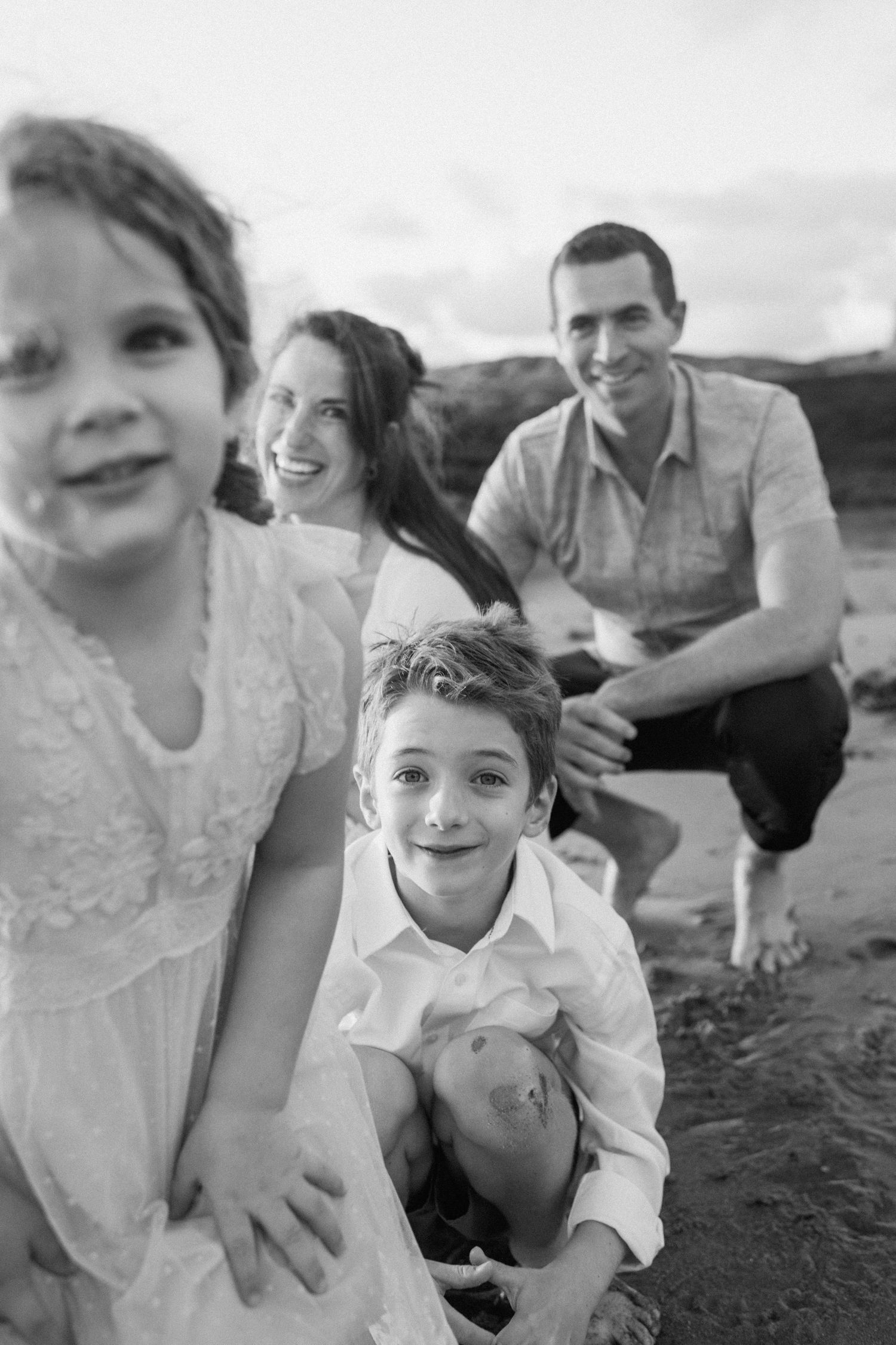 Black and white beach photo of smiling family with two young children crouching in sand.