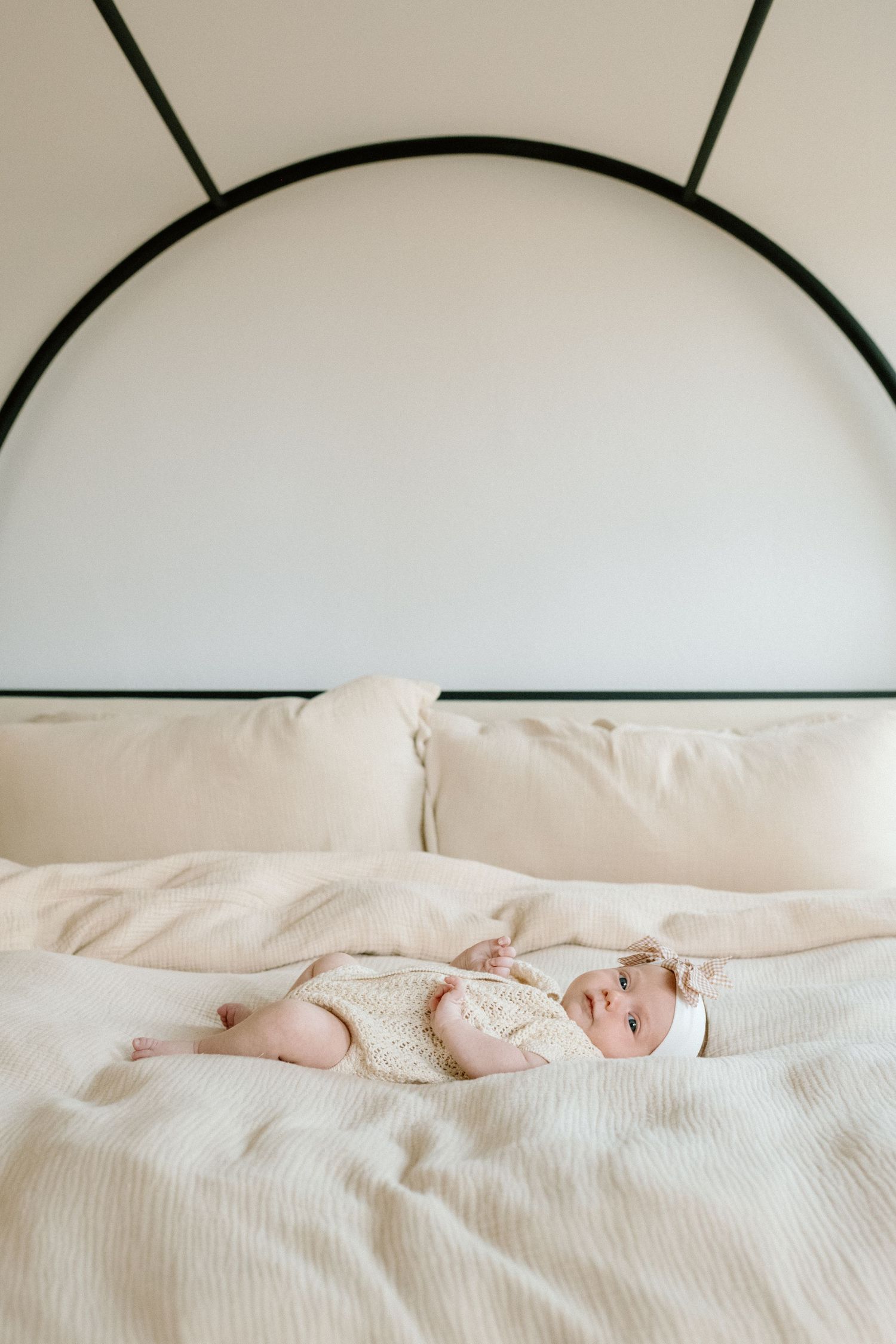 Newborn baby in cream knit outfit lying on a white bed with arched headboard.