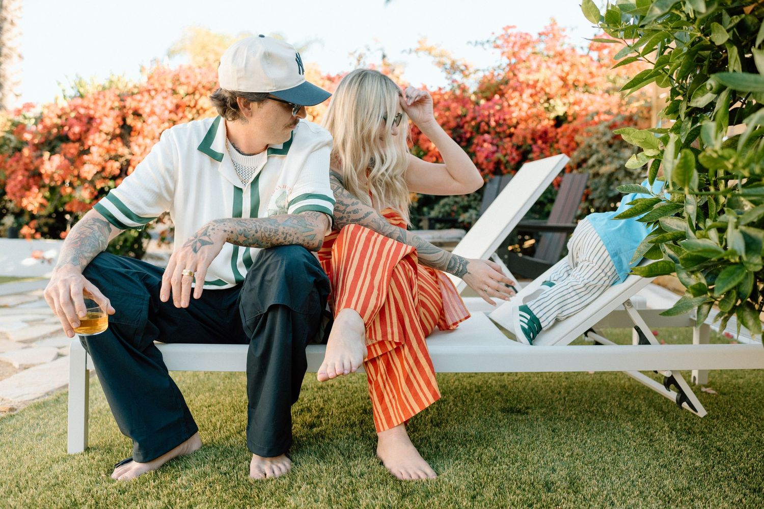 Two people relax on leisure chairs in a backyard garden setting with autumn foliage in the background.