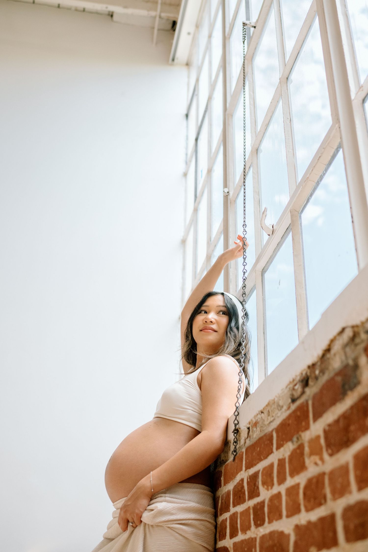 Pregnant woman leans against wall with arm up during maternity photoshoot in a San Diego studio.