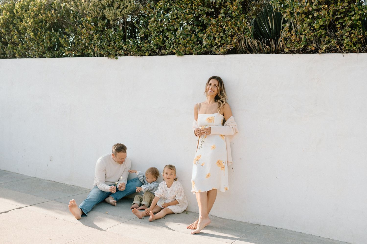 Family of four posing against white wall outdoors, mother in floral dress holding baby, father sitting with toddler.