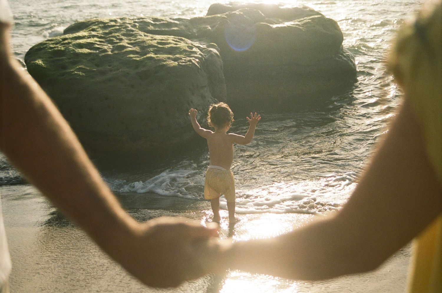 Child playing at beach shoreline framed by parents holding hands, with sunlit rocks and ocean waves in background.