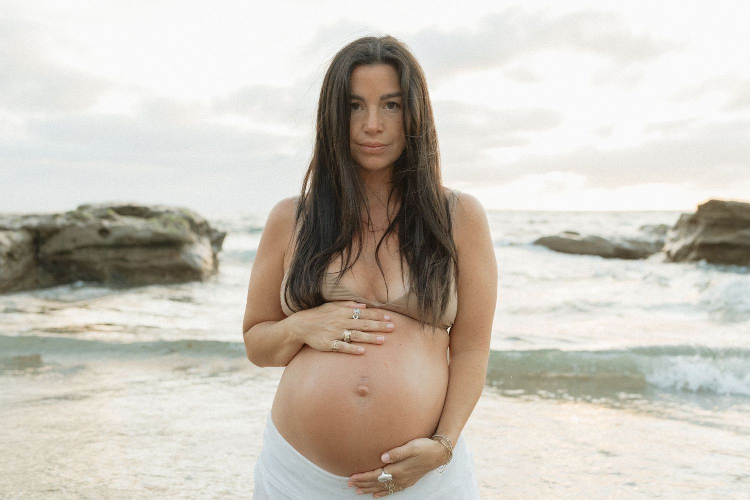 A pregnant woman in a white wrap cradles her bare belly while standing on a rocky beach at sunset.