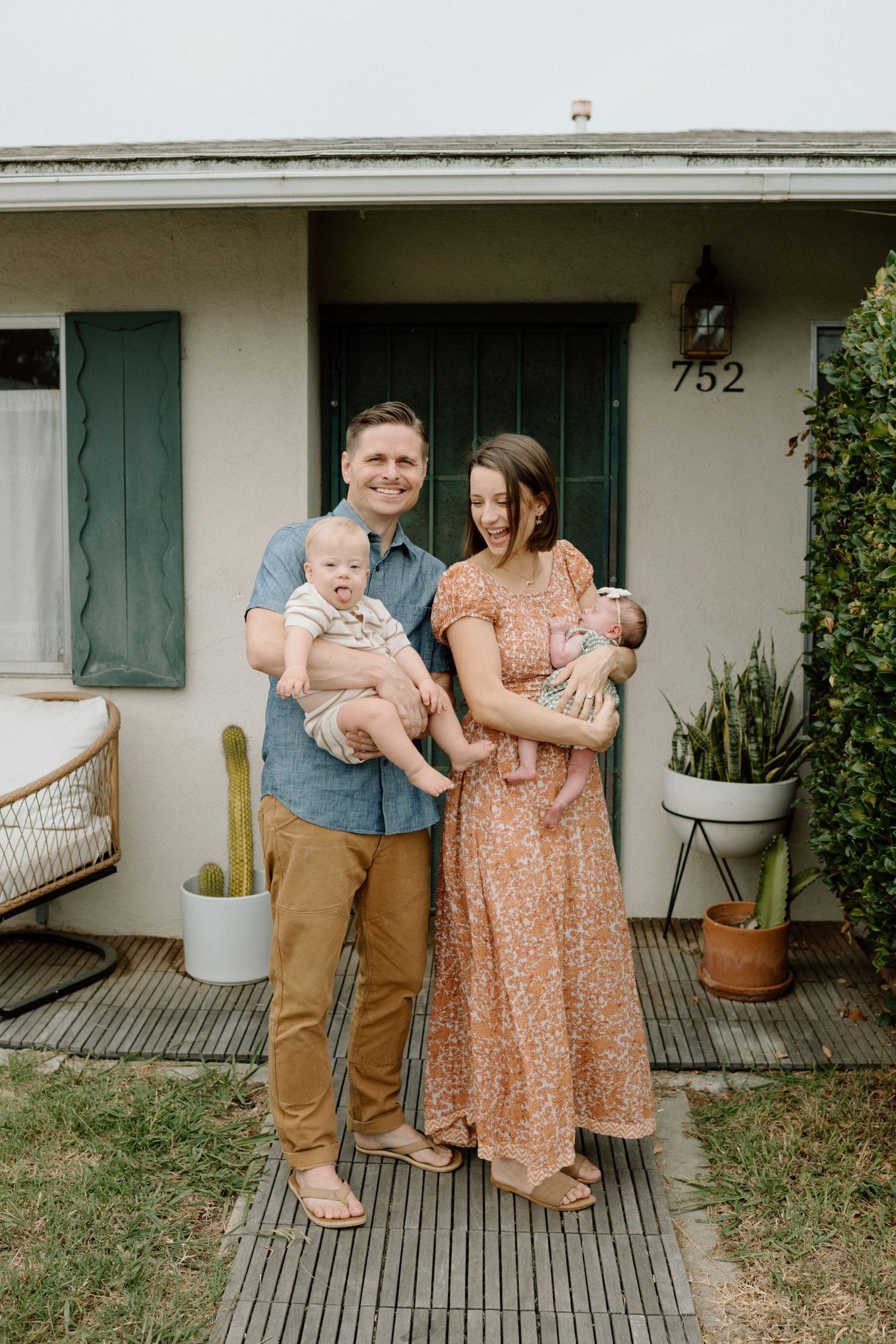 Smiling couple stands in front of their home, each holding a young baby outdoors.