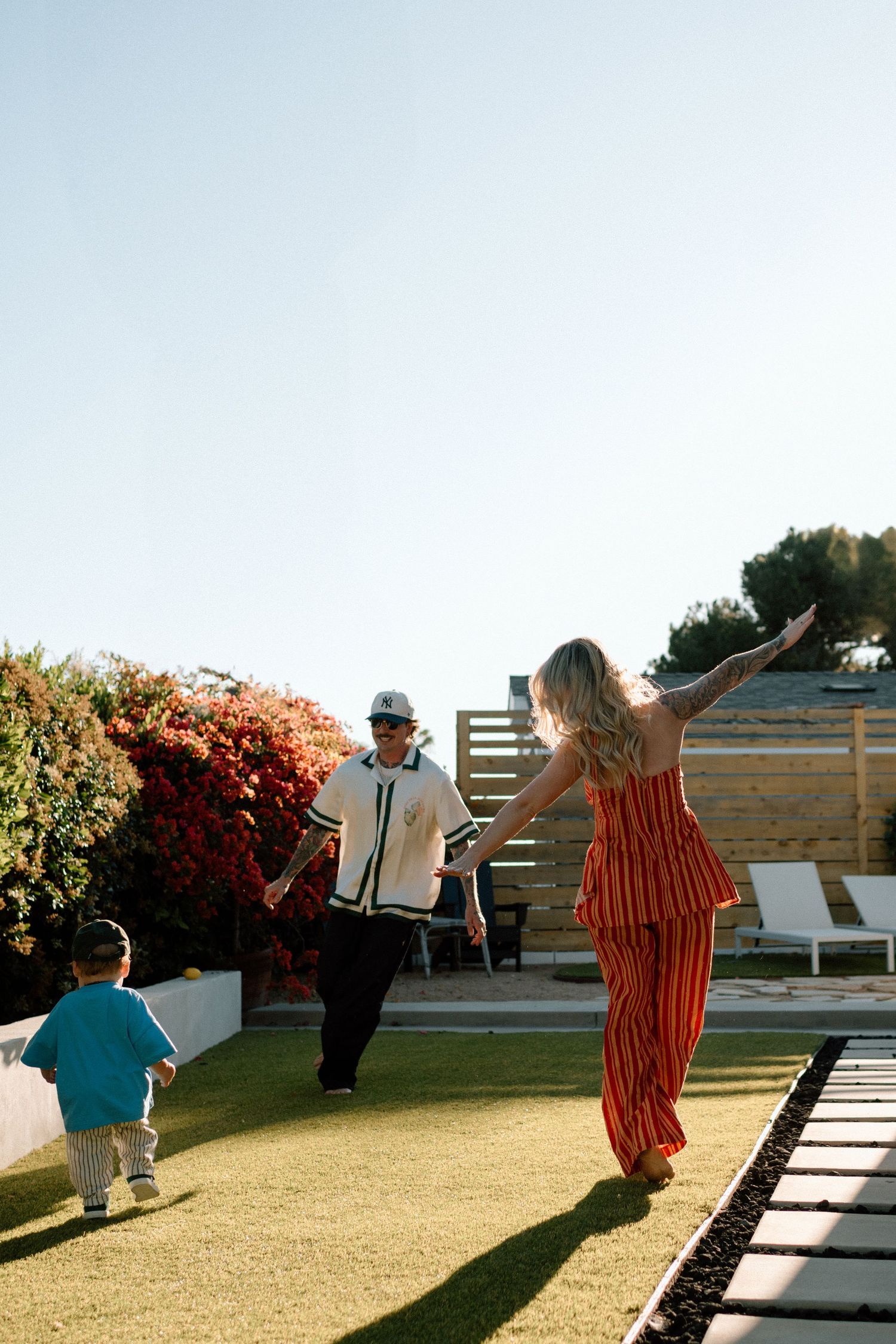 Family plays together on a sunny day in a backyard with artificial turf and garden features.
