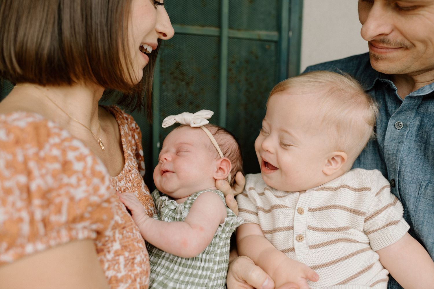 Happy family with parents holding a laughing toddler and newborn baby in green outfit indoors.