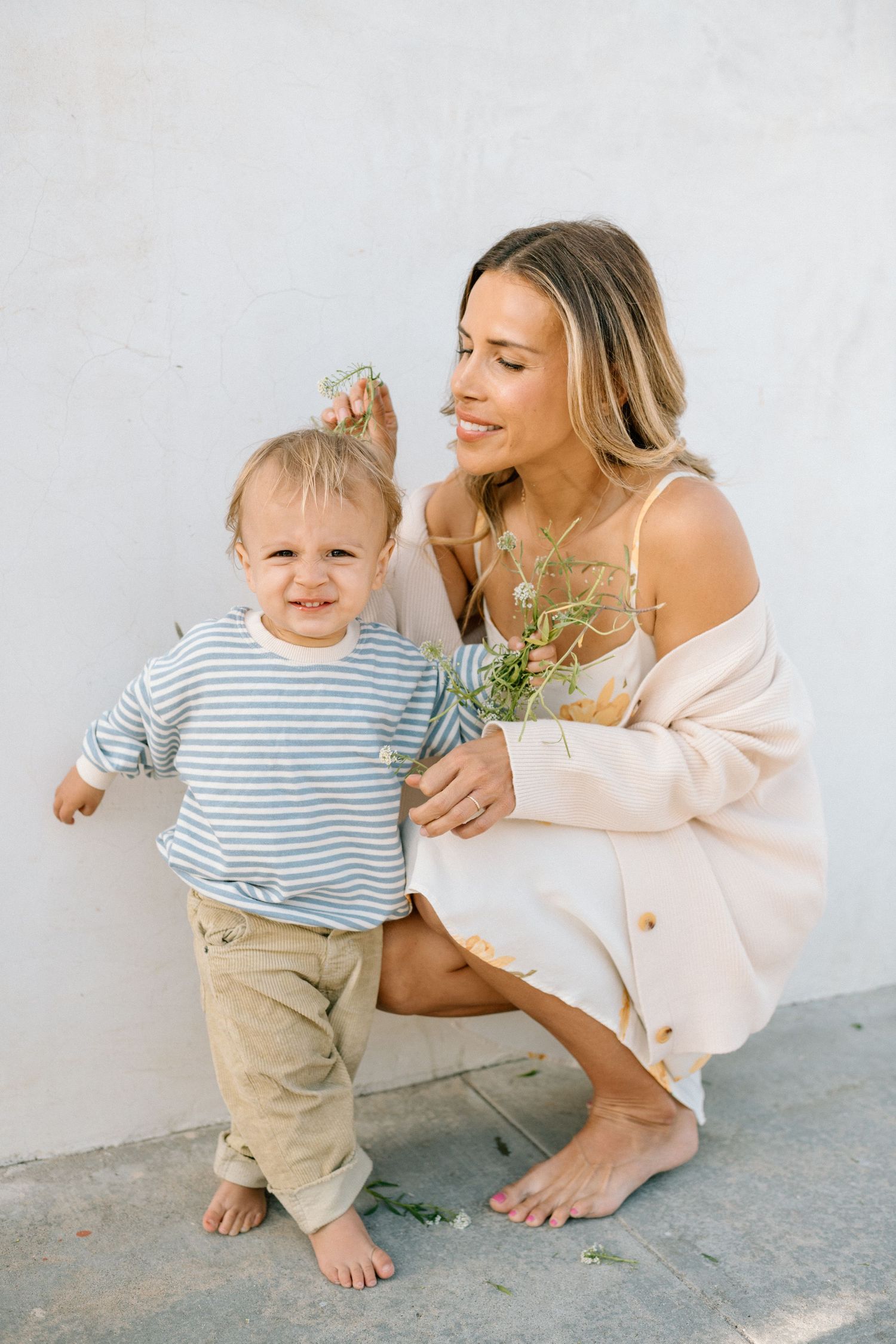 A smiling mother crouches beside her toddler near a white wall, both barefoot, as she holds small wildflowers.