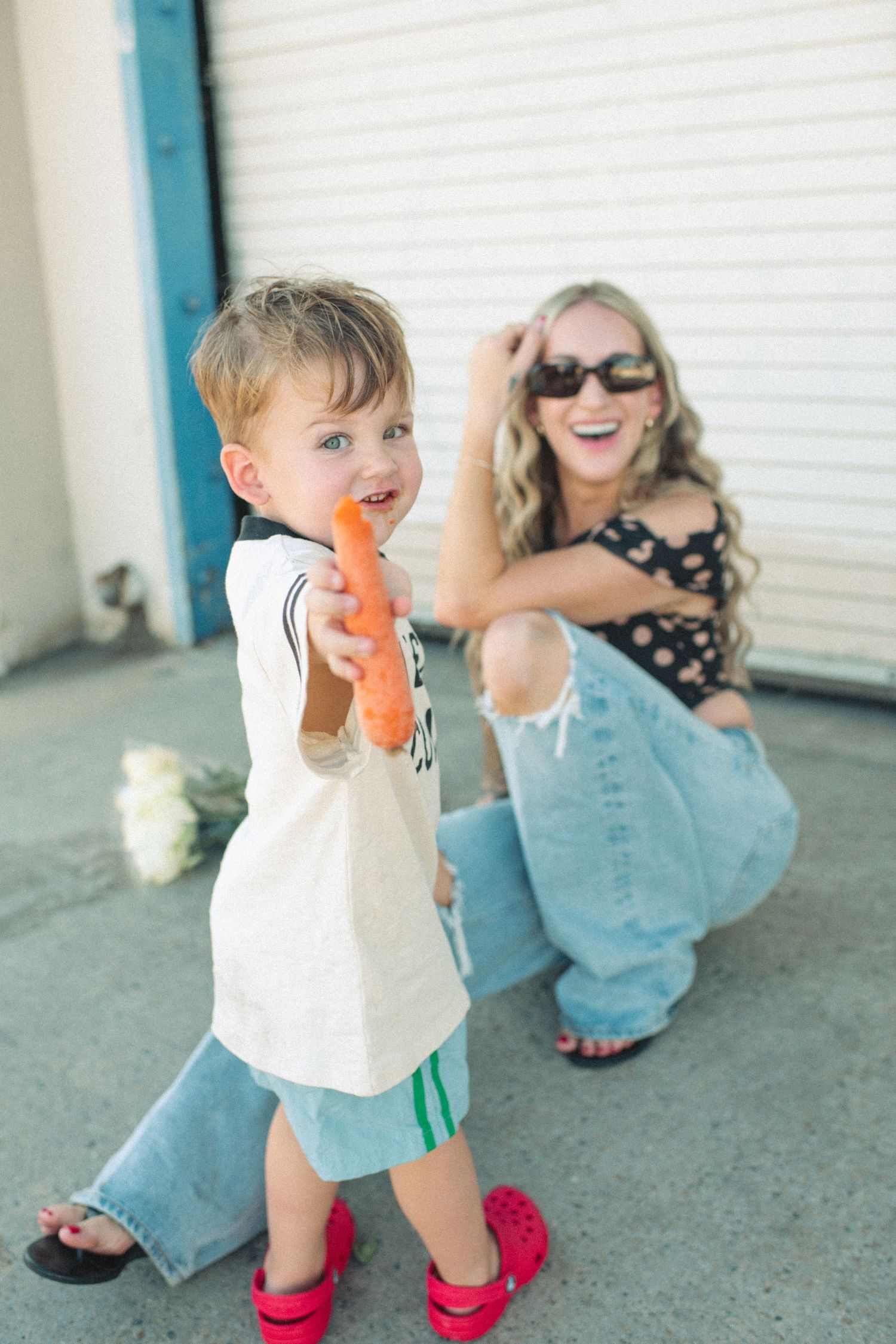 Toddler boy holding a carrot toward camera while blonde mom in sunglasses laughs behind him outdoors.