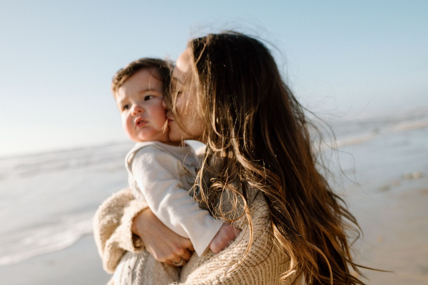 Mother kissing baby on cheek at sunny beach with ocean backdrop.