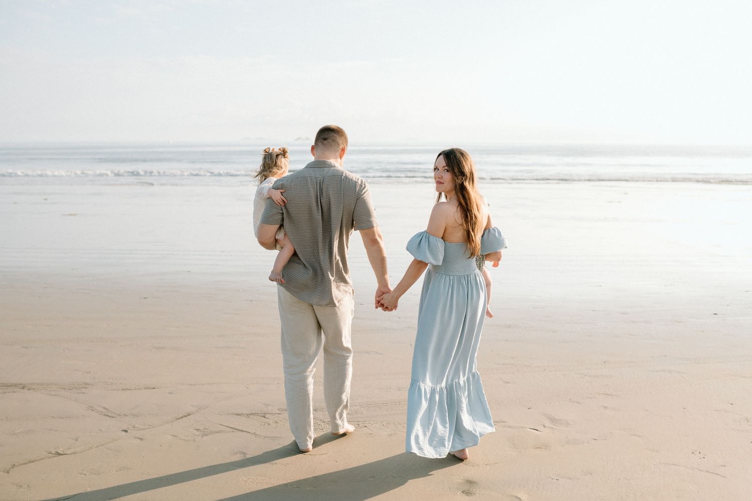 Couple holding hands walking toward ocean waves with baby at beach.