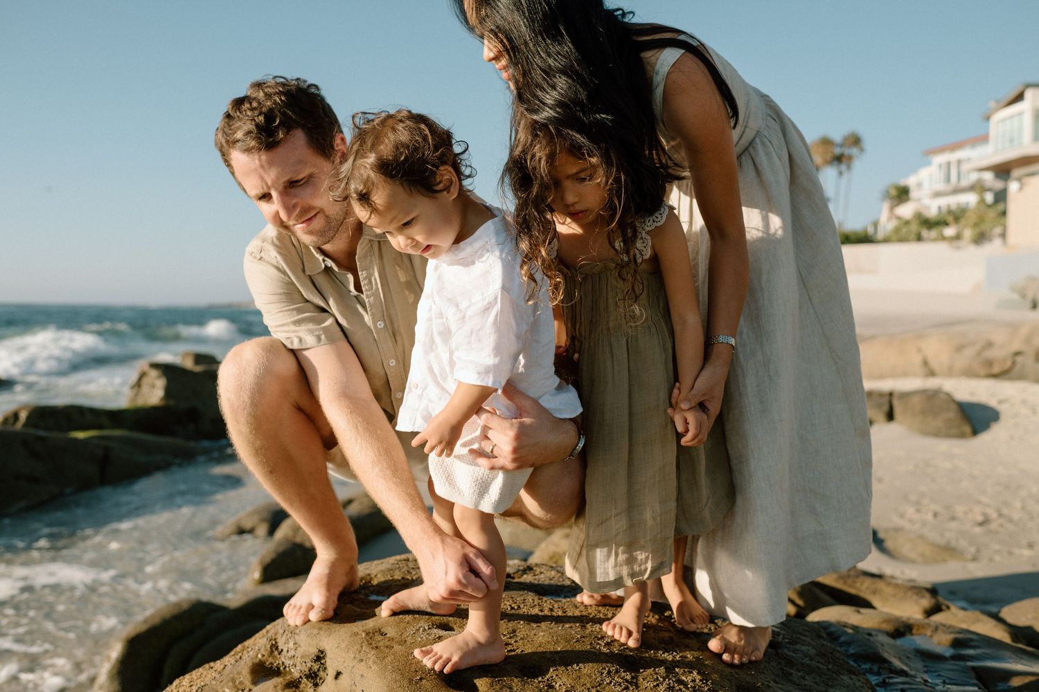A family of four explores tide pools together on a sunny rocky beach coastline.