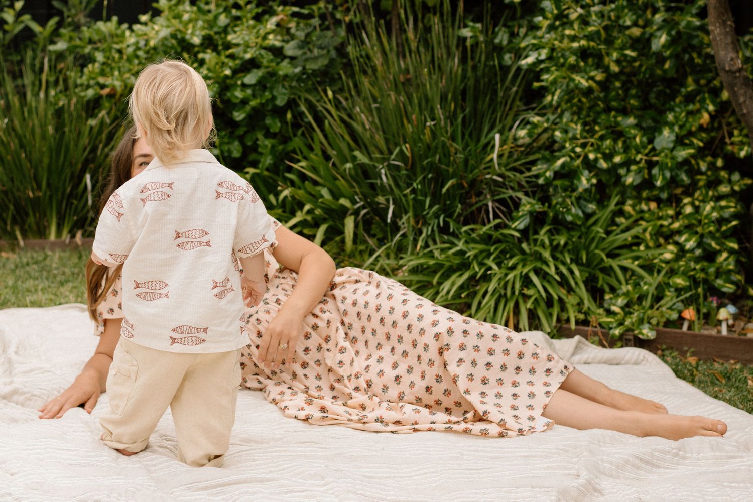 Young child sits beside pregnant mother resting on a blanket in a garden setting.