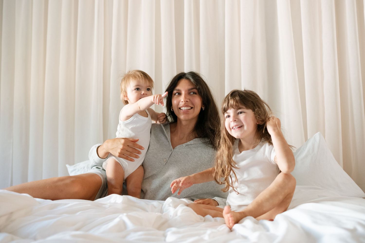 A smiling mother sits on a white bed holding a baby while her young daughter leans against her, all in casual white outfits.