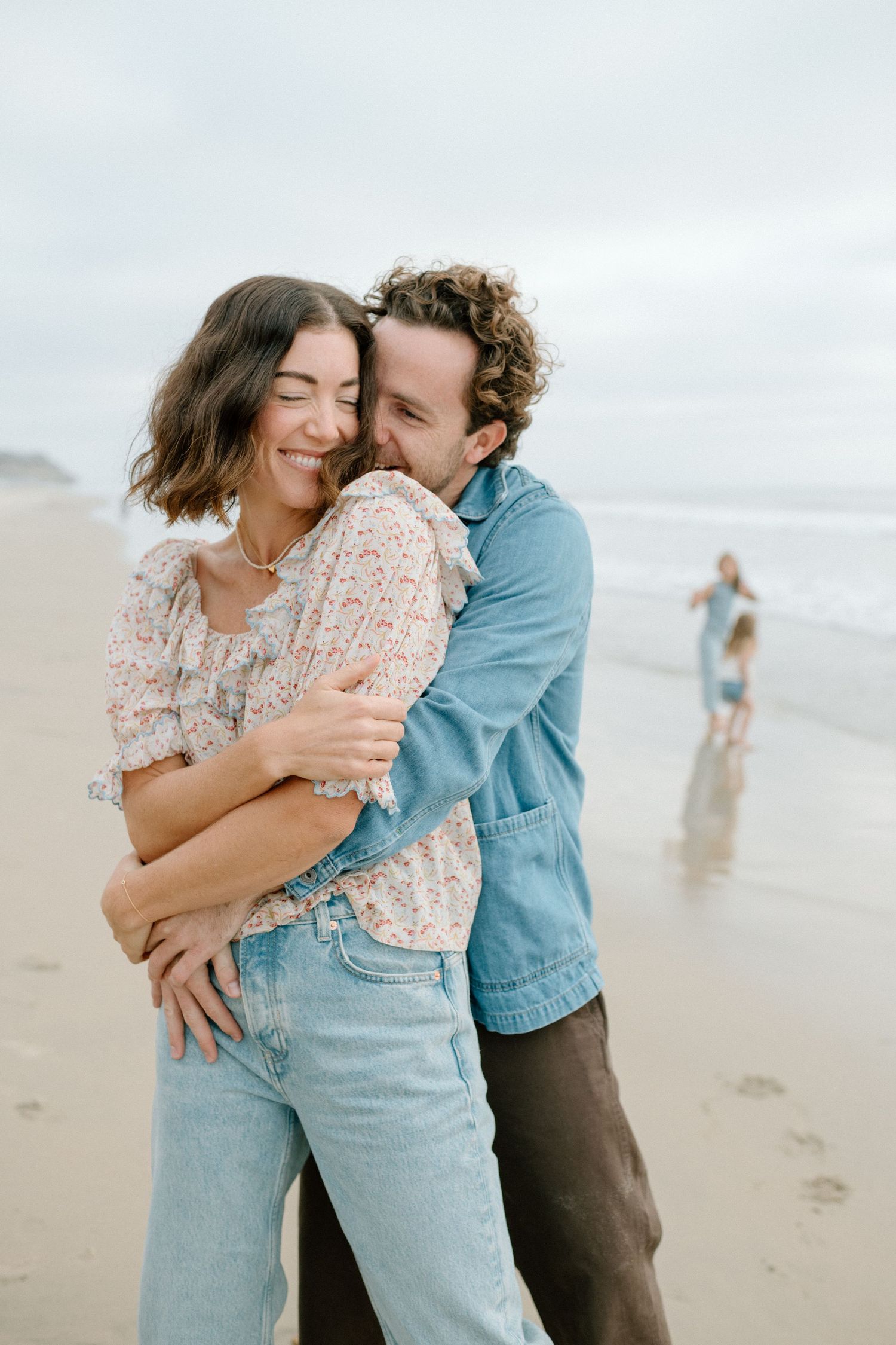 Man embraces woman from behind as they smile together on a beach with a child playing nearby.