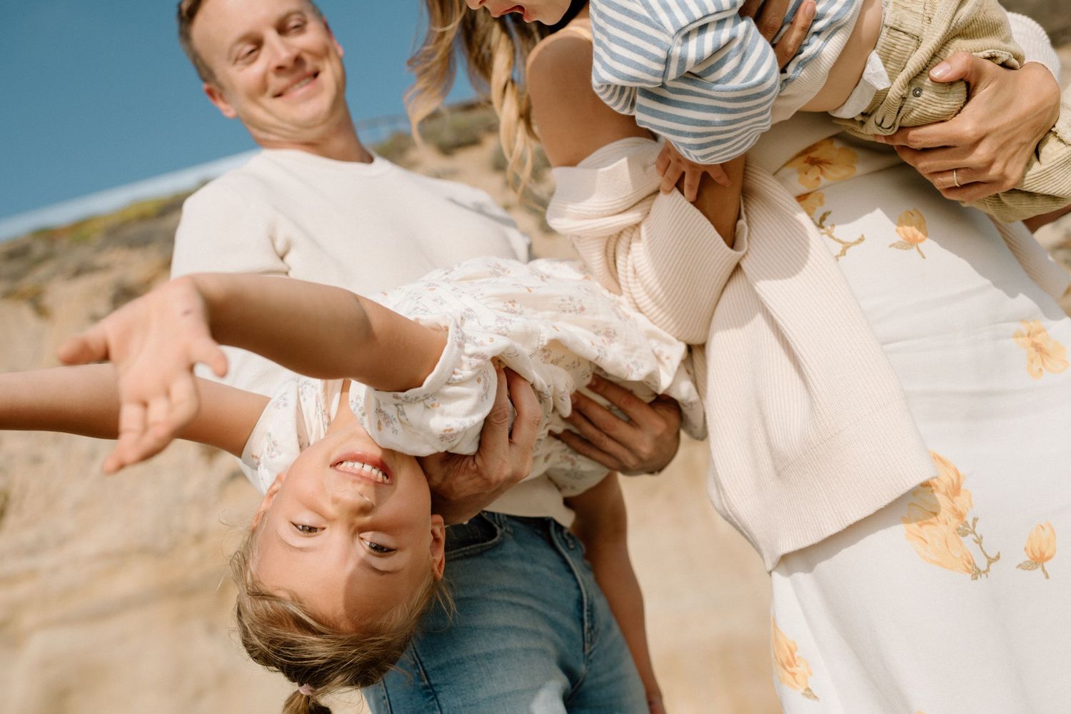 Happy family playing outdoors on a sunny beach, parents lifting laughing child upside down against blue sky.