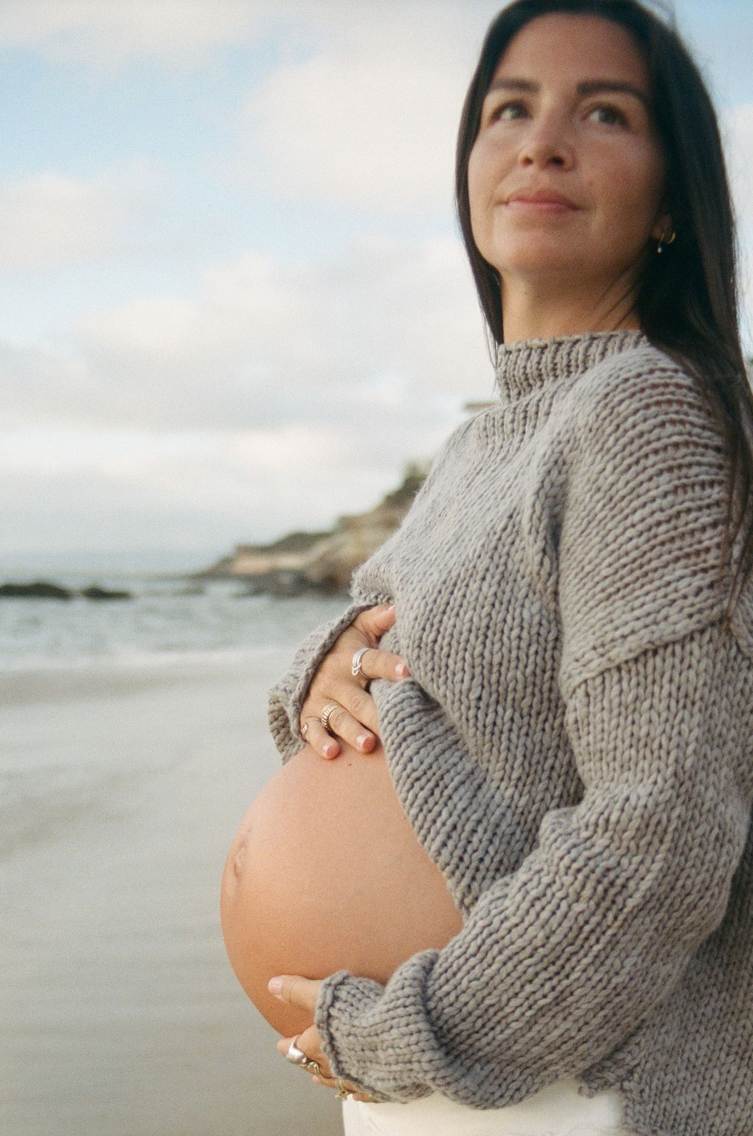 Pregnant woman in gray knit sweater cradling baby bump on a scenic beach, gazing upward with a serene expression.