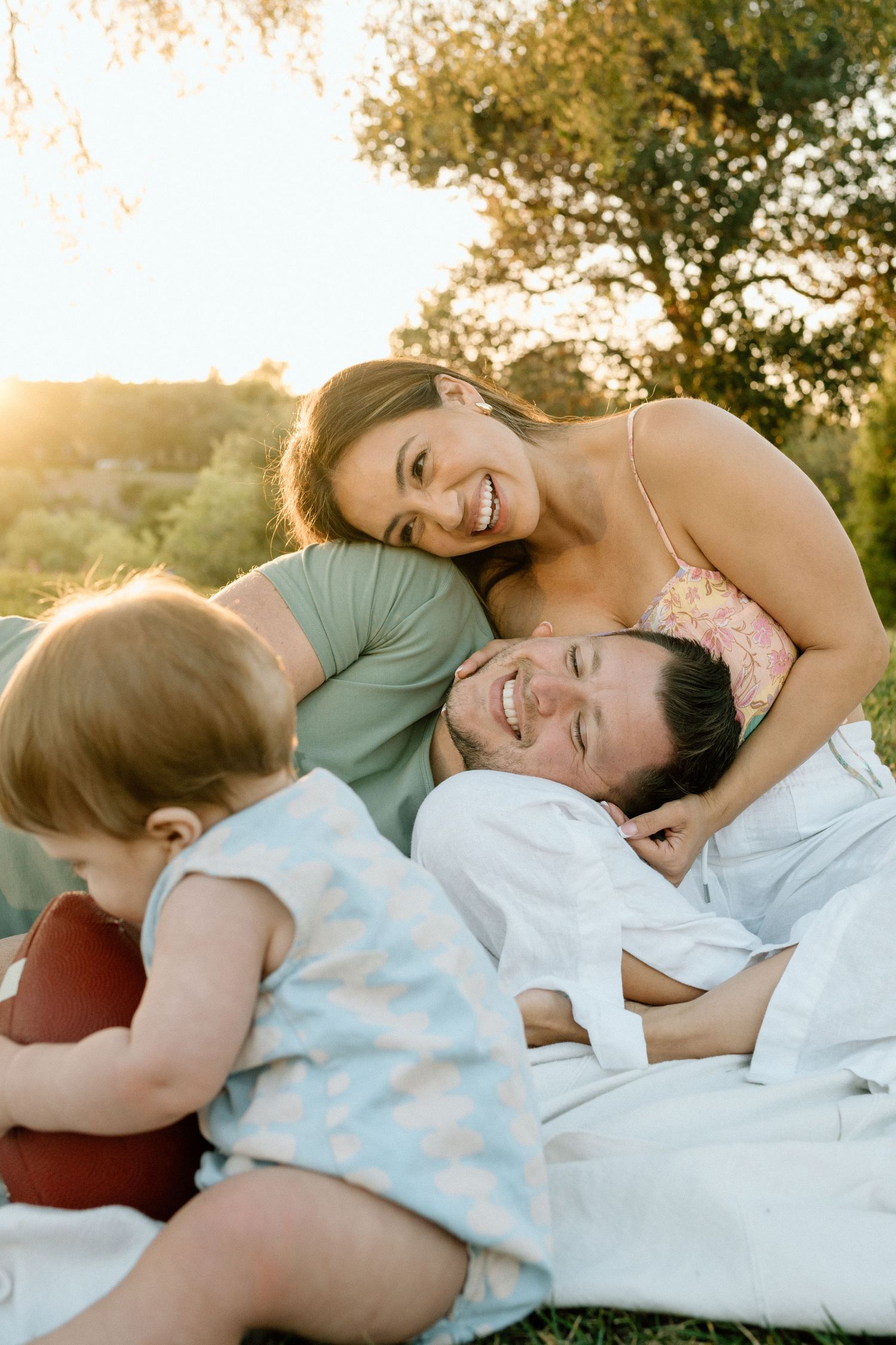 Happy family relaxing outdoors on a sunny day, with parents smiling and a toddler playing on a blanket in golden hour light.