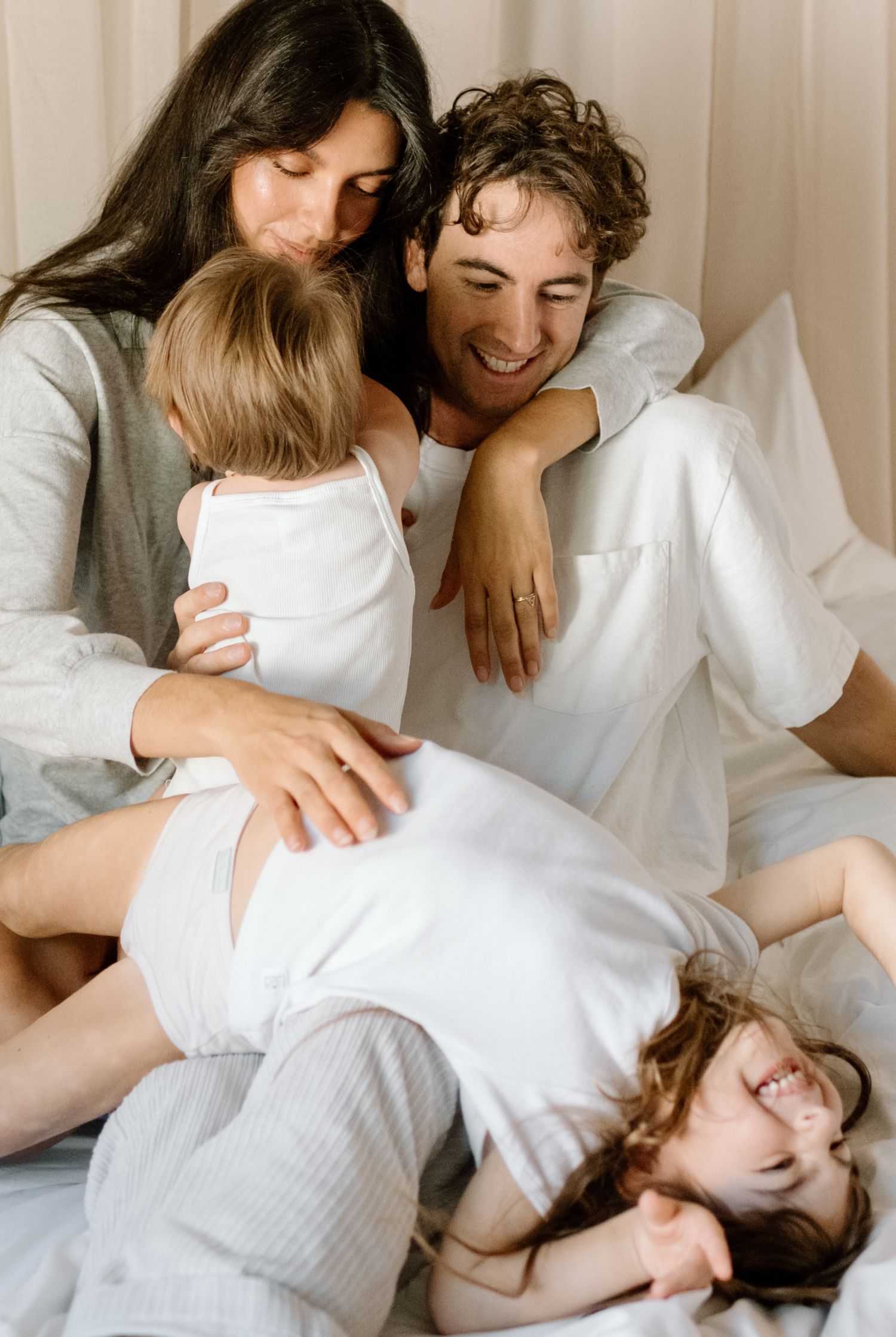 Happy family with two young children laughing and cuddling together on a white bed in matching white outfits.