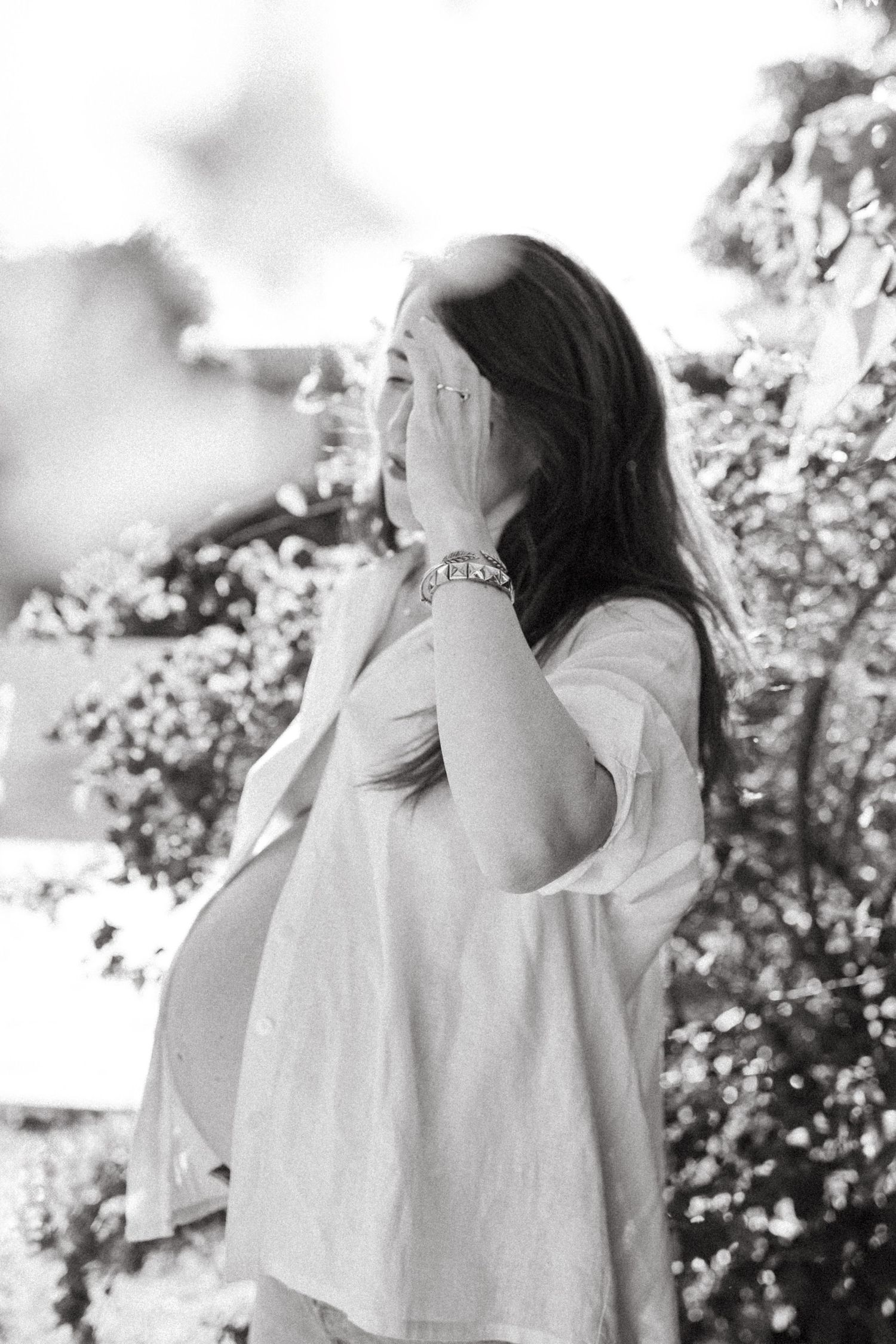 Black and white portrait of pregnant woman covering face with hand among blooming flowers.
