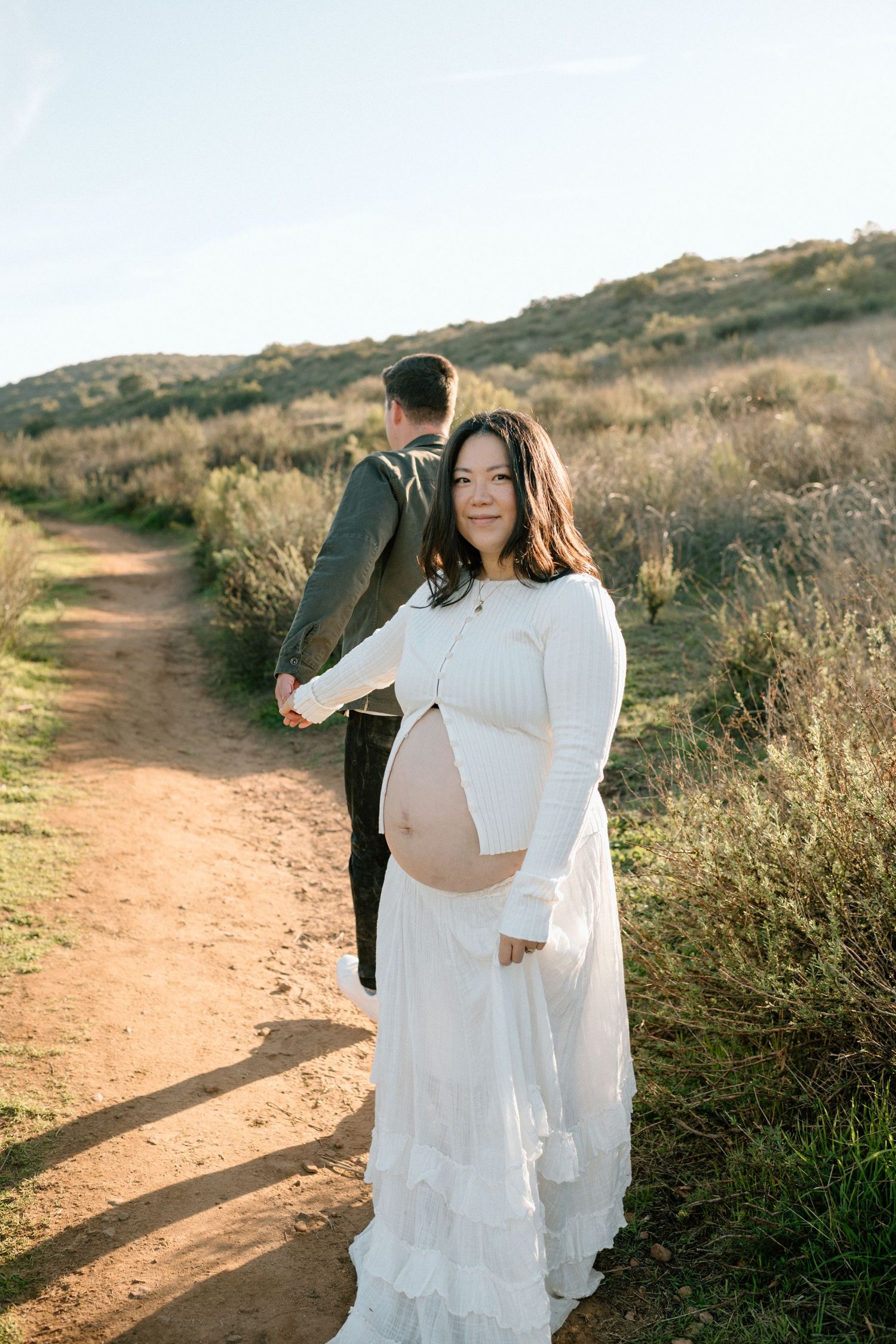 Pregnant woman in white lace dress holds partners hand on sunny dirt trail maternity photoshoot.