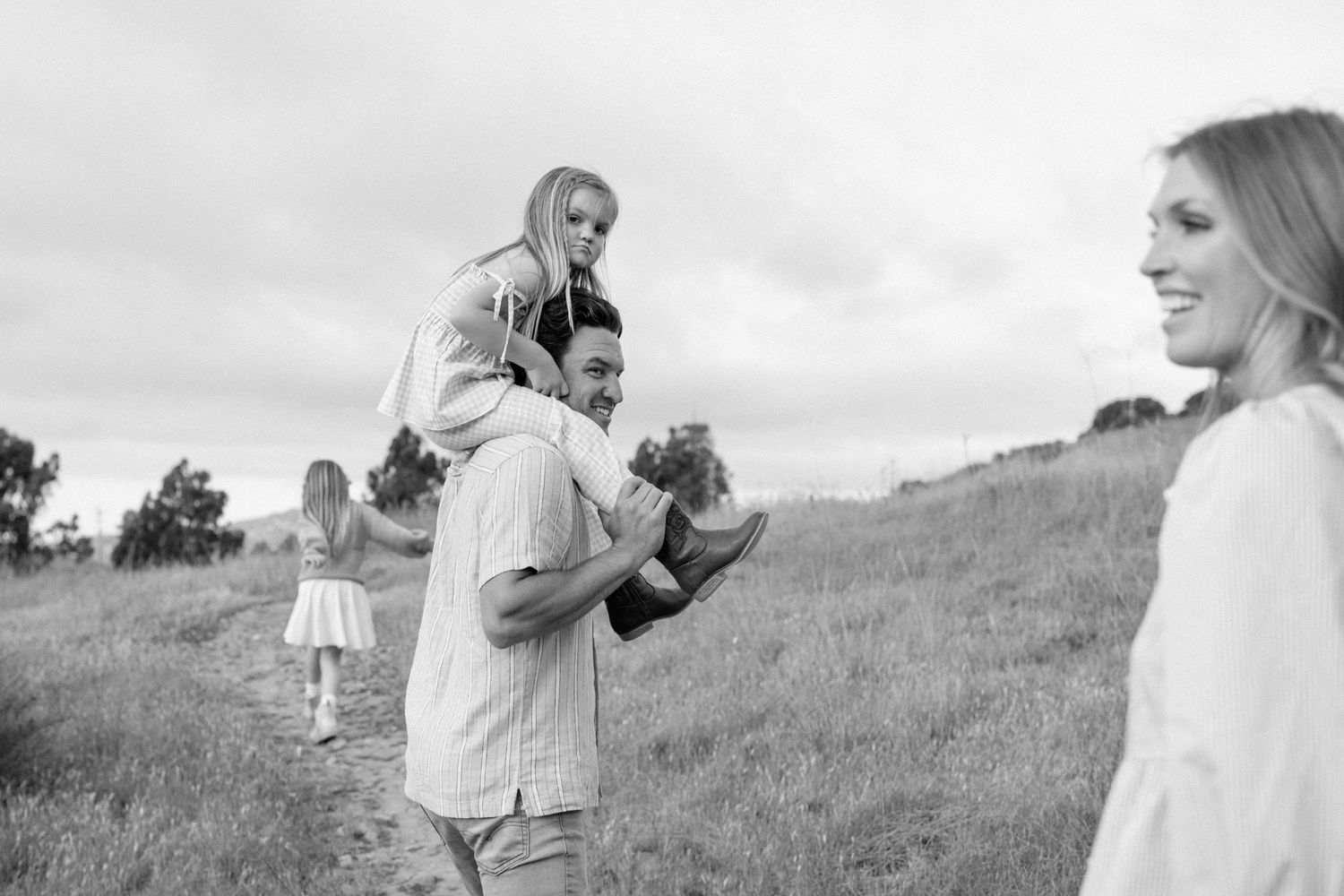Black and white photo of a father carrying his daughter on his shoulders while walking through a grassy hillside path.