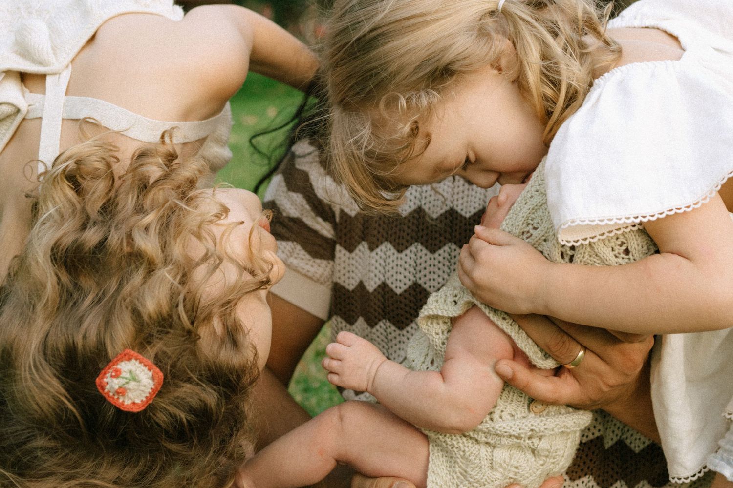 Two young girls lean over a baby held by an adult outdoors in a warm, candid family moment.
