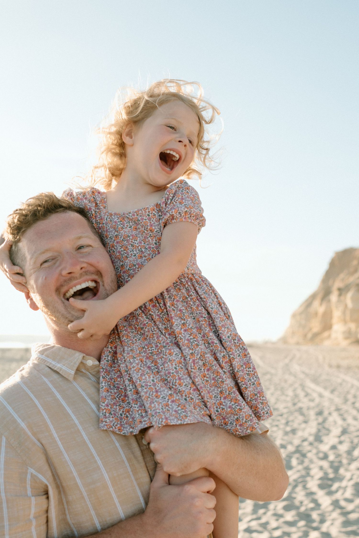 Laughing father holds his giggling daughter on his shoulders on a sunny sandy beach.