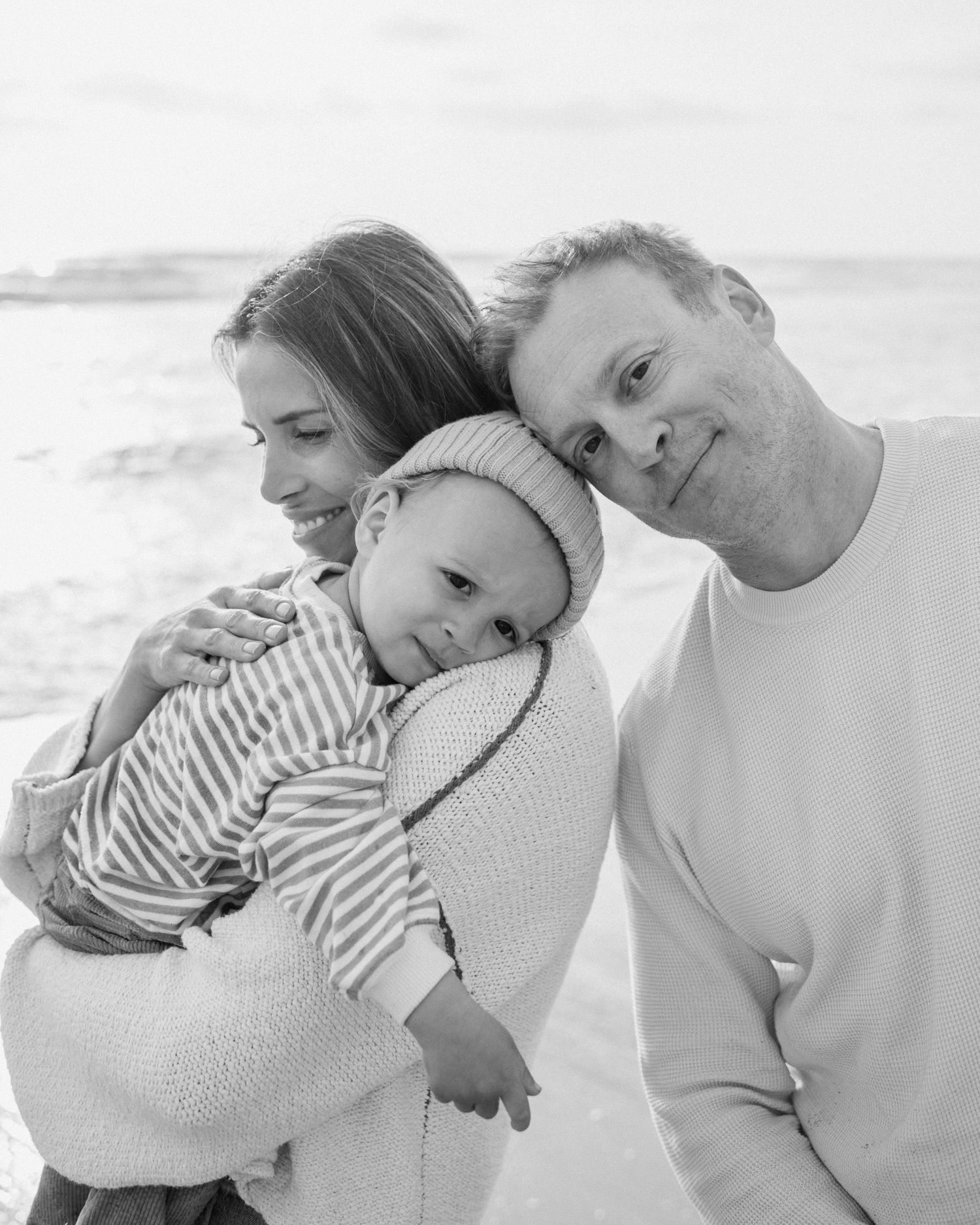 A smiling couple cuddles their newborn baby wrapped in a knit blanket on a beach in black and white.