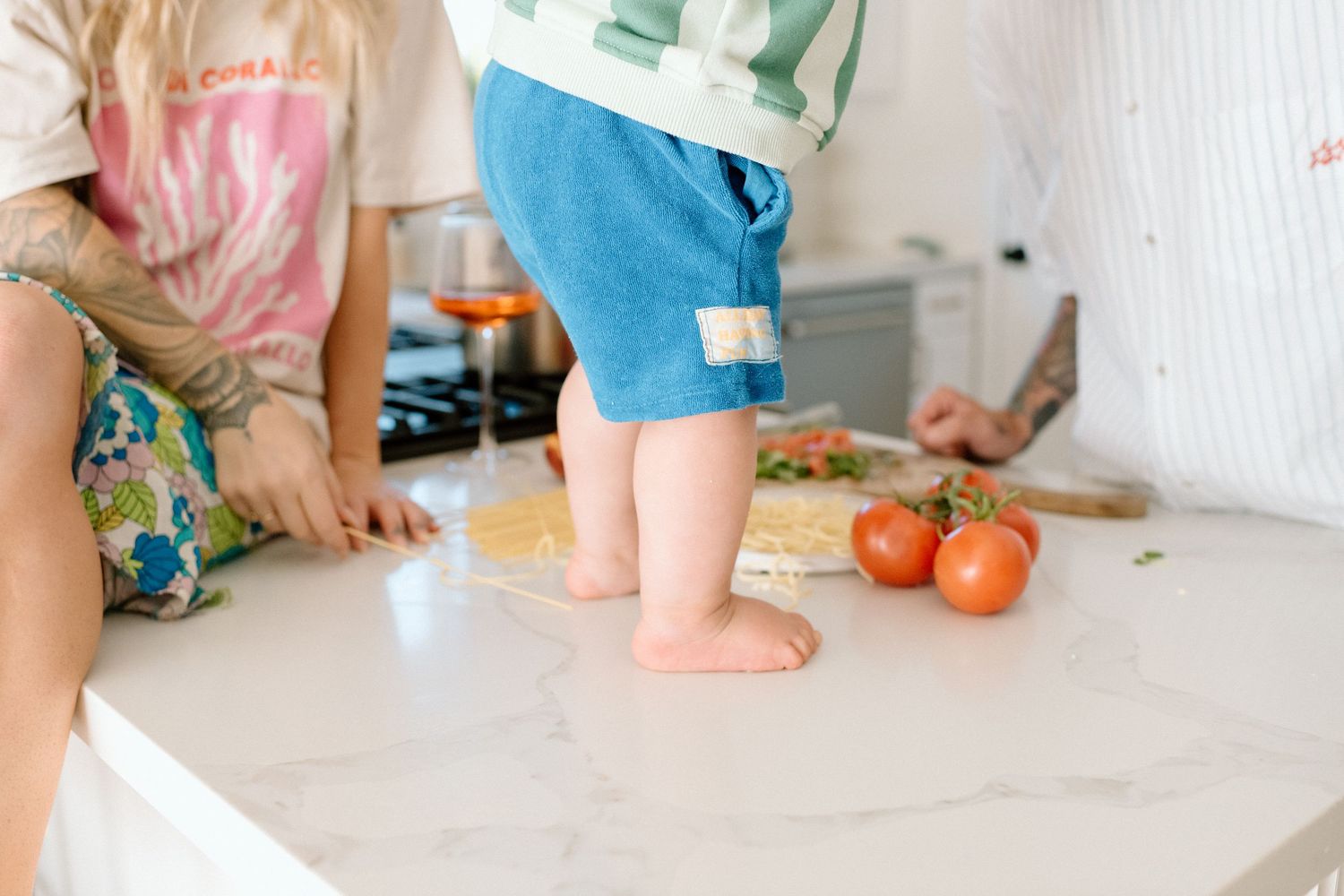 Small children's bare feet visible on white kitchen counter with fresh tomatoes while meal preparation is happening.